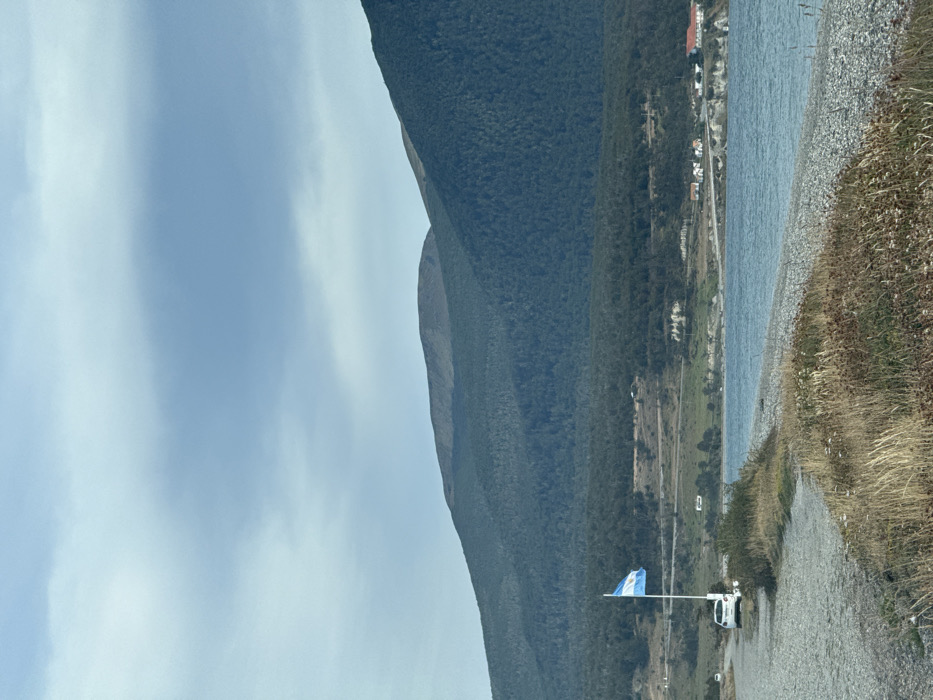 The image displays a landscape in the Municipio de Tolhuin, Argentina, under an overcast, light blue-grey sky. In the background, a range of large, dark green, densely forested mountains dominates, with higher elevations appearing less vegetated. The midground features a body of water, likely a lake or bay, with a visible pebble beach along its shore to the right. Across the water and further back, a small settlement is discernible, comprising several buildings, including structures with white walls and red roofs, situated on a slightly elevated terrain with a road running nearby. In the left midground, a gravel path extends parallel to the water's edge. An Argentinian flag, consisting of blue and white horizontal stripes, flies from a white flagpole positioned to the left of the path. A white passenger vehicle is parked on the gravel path behind the flagpole. The immediate foreground is characterized by sparse, dry golden-brown and green grasses and weeds. No individuals are visible.