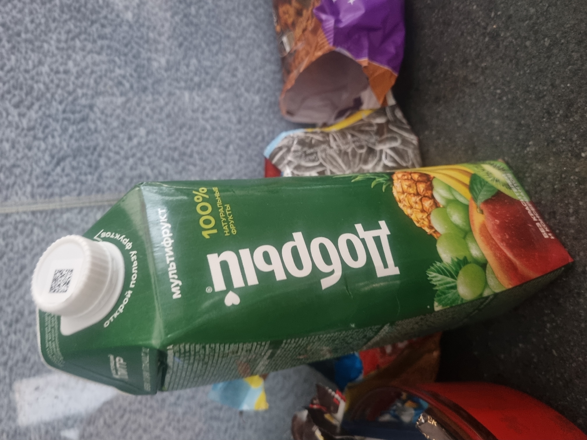 A close-up, slightly angled shot captures a carton of juice with the brand name "Добрый" (Dobry) prominently displayed. The carton is green and features an illustration of various fruits, including pineapple, grapes, bananas, and what appears to be a peach or nectarine. Above the fruit illustration, the text "МУЛЬТИФРУКТ" (Multifruit) is visible, along with "100% НАТУРАЛЬНЫЕ ФРУКТЫ" (100% Natural Fruits). A white cap with a QR code is on top of the carton. The carton is placed on a dark, speckled surface, possibly a countertop or table. In the background, out-of-focus, are several crumpled food packaging items, one appearing to be a bag of sunflower seeds and another a purple and orange package. The overall environment appears to be indoors with a textured, gray wall in the background. The specific location in Samara, Russia cannot be determined from the visual information provided.