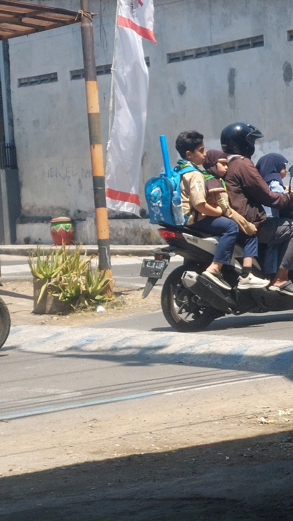 In Sidoarjo, Indonesia, a family of four rides a motorcycle. The parents are seated in the front and middle, with two children riding behind them. The children appear to be young school-aged boys. The boy in the middle wears a scout uniform and carries a blue backpack. The motorcycle is black and silver. The family is on a paved road, possibly a street or a highway, with a pedestrian crossing marked by white lines. In the background, a building with a gray concrete wall and a flag pole with a white flag are visible. The environment is bright and sunny, suggesting daytime. The overall scene depicts a common mode of transportation in the region and a snapshot of daily life.
