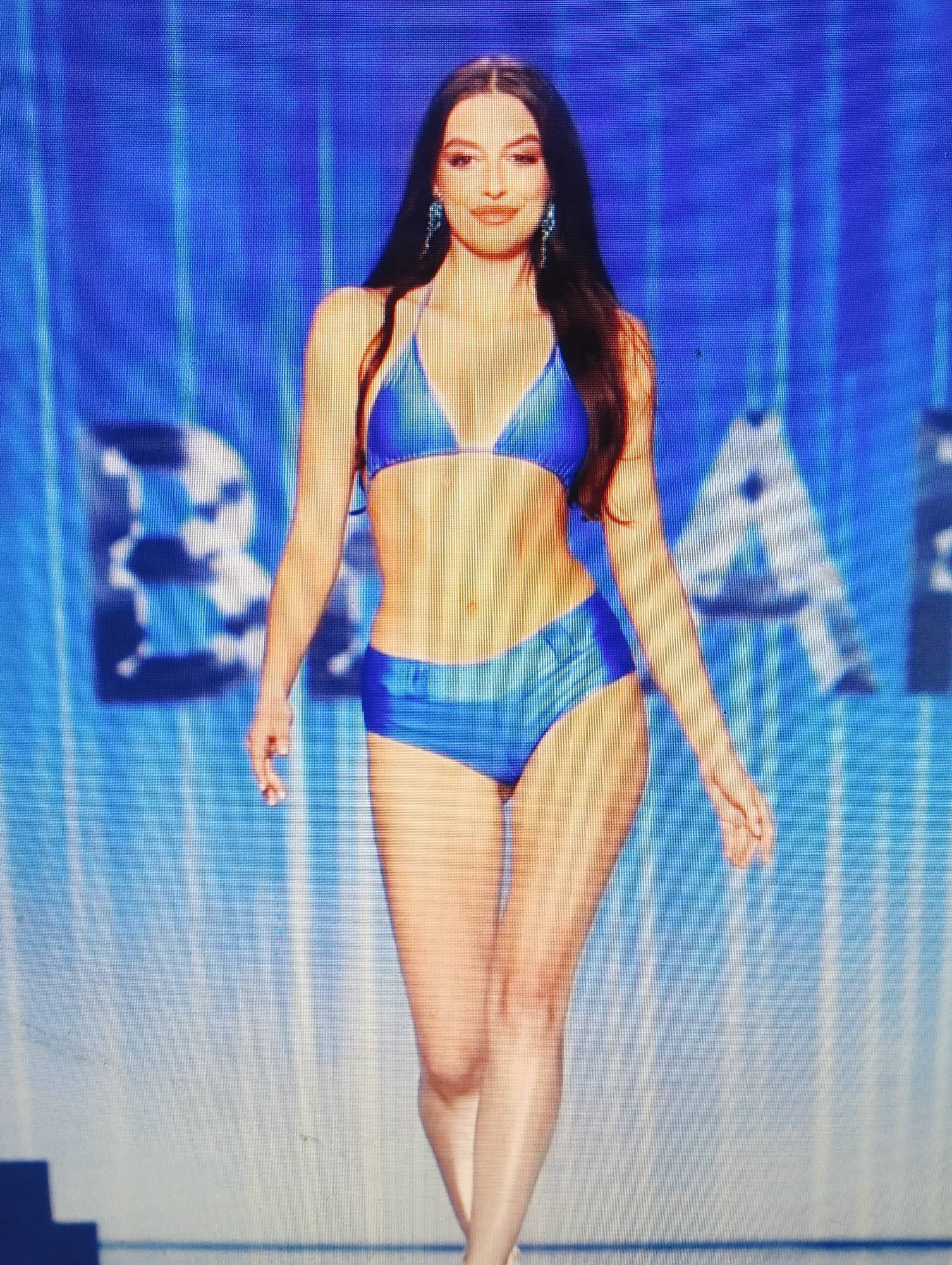 A young woman with long dark hair and wearing a blue bikini walks across a stage. She has on long dangling earrings and is walking towards the viewer. The background is a bright blue screen with vertical lines. Large white letters are visible in the background, which appear to be part of a word or phrase. The floor of the stage is light gray.