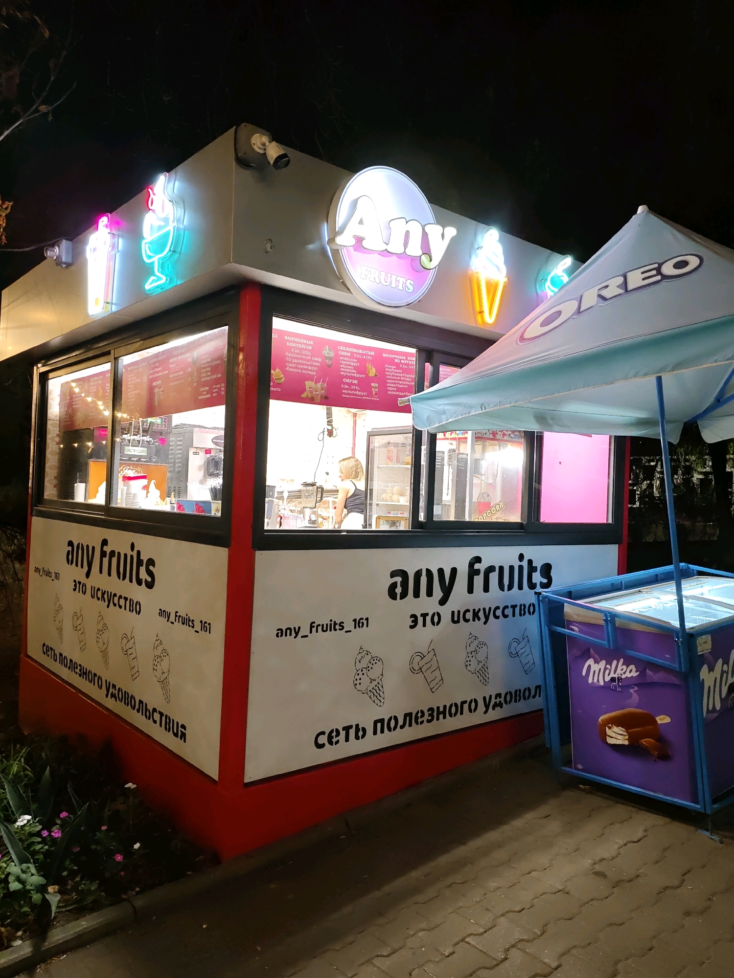 This image depicts an outdoor ice cream and fruit drink stand operating at night in the city of Rostov-on-Don, Russia. The stand features a prominent neon sign that reads "Any FRUITS," and other neon lights shaped like ice cream cones and drinks. A menu is visible behind a glass window, displaying various items in Russian. Inside the stand, a woman appears to be preparing a beverage. Adjacent to the stand, a blue umbrella with the "OREO" logo is set up, and a freezer unit branded with "Milka" is visible, containing ice cream products. The ground is paved with interlocking bricks, and some greenery is present to the left of the stand. The lighting suggests it is nighttime.