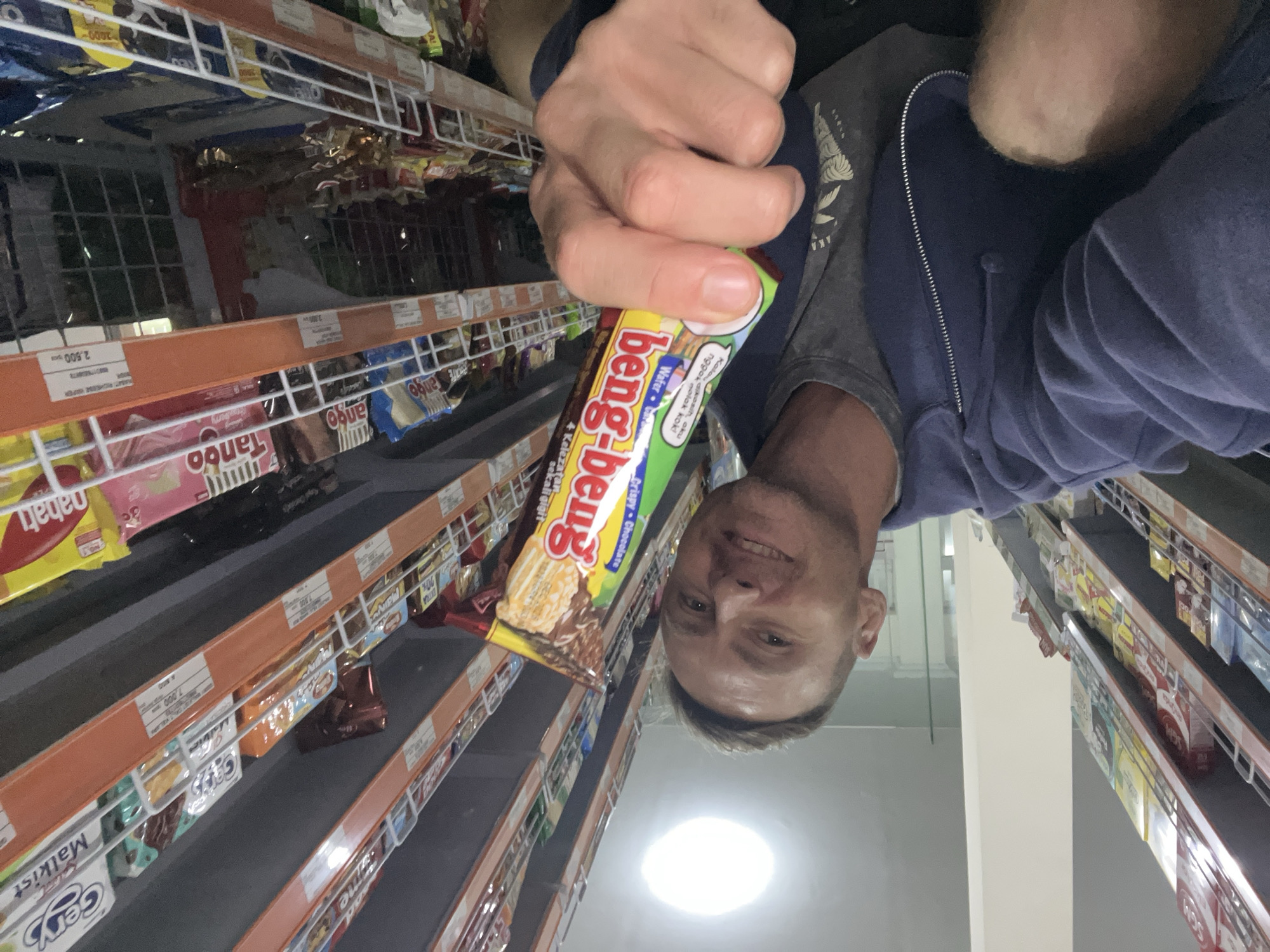 A man is holding a Beng-Beng wafer in a grocery store, likely in Sukawati, Indonesia based on the "Beng-Beng" brand and other Indonesian snack brands visible on the shelves such as "Tango" and "Nabati". The shelves are stocked with various snacks and candies. The image is taken from an unusual angle, looking up at the man. He is wearing a grey t-shirt and a blue zip-up hoodie. Price tags are visible on the shelves.