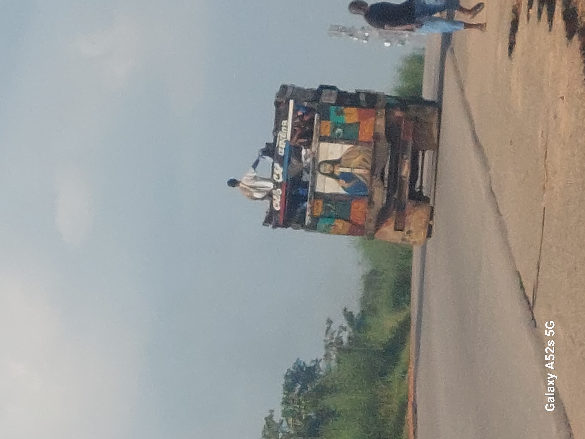 In Awkuzu, Nigeria, a truck is seen on a road with greenery in the background. The truck is decorated with a painting of Jesus on one side and has a person standing on top of it. Another person is walking on the side of the road. The sky is overcast.