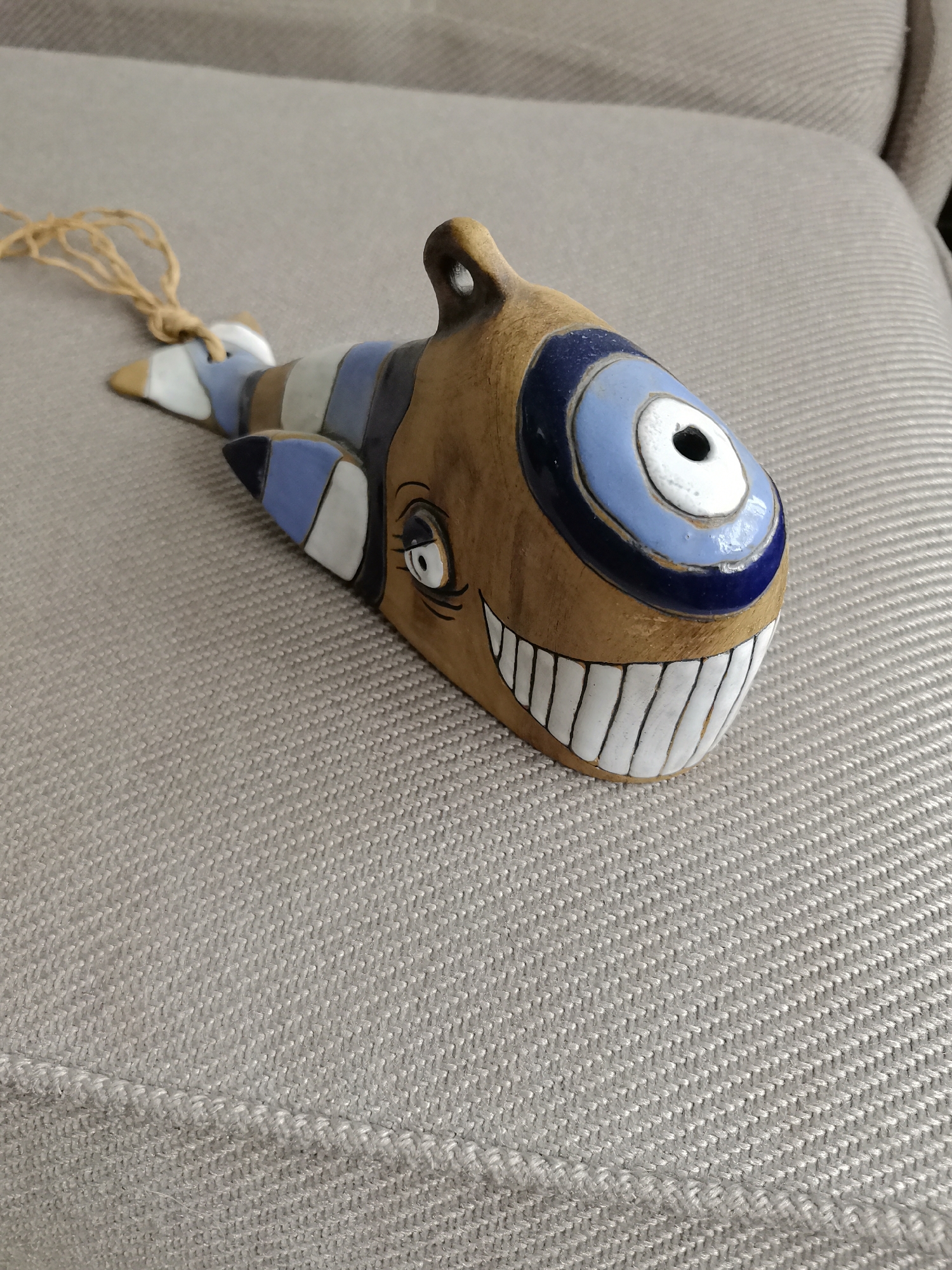 A wooden fish ornament with painted details is lying on a textured, light-colored surface. The fish is brown with blue, white, and gray stripes along its body. It has a large blue and white eye and a wide, smiling mouth with white teeth. A loop is at the top of the fish's head, with a thin, light-brown string attached to it, hanging to the left. The background is out of focus and appears to be upholstery with a woven texture. There is no indication of the city of Oryol, Russia, or any other specific location.