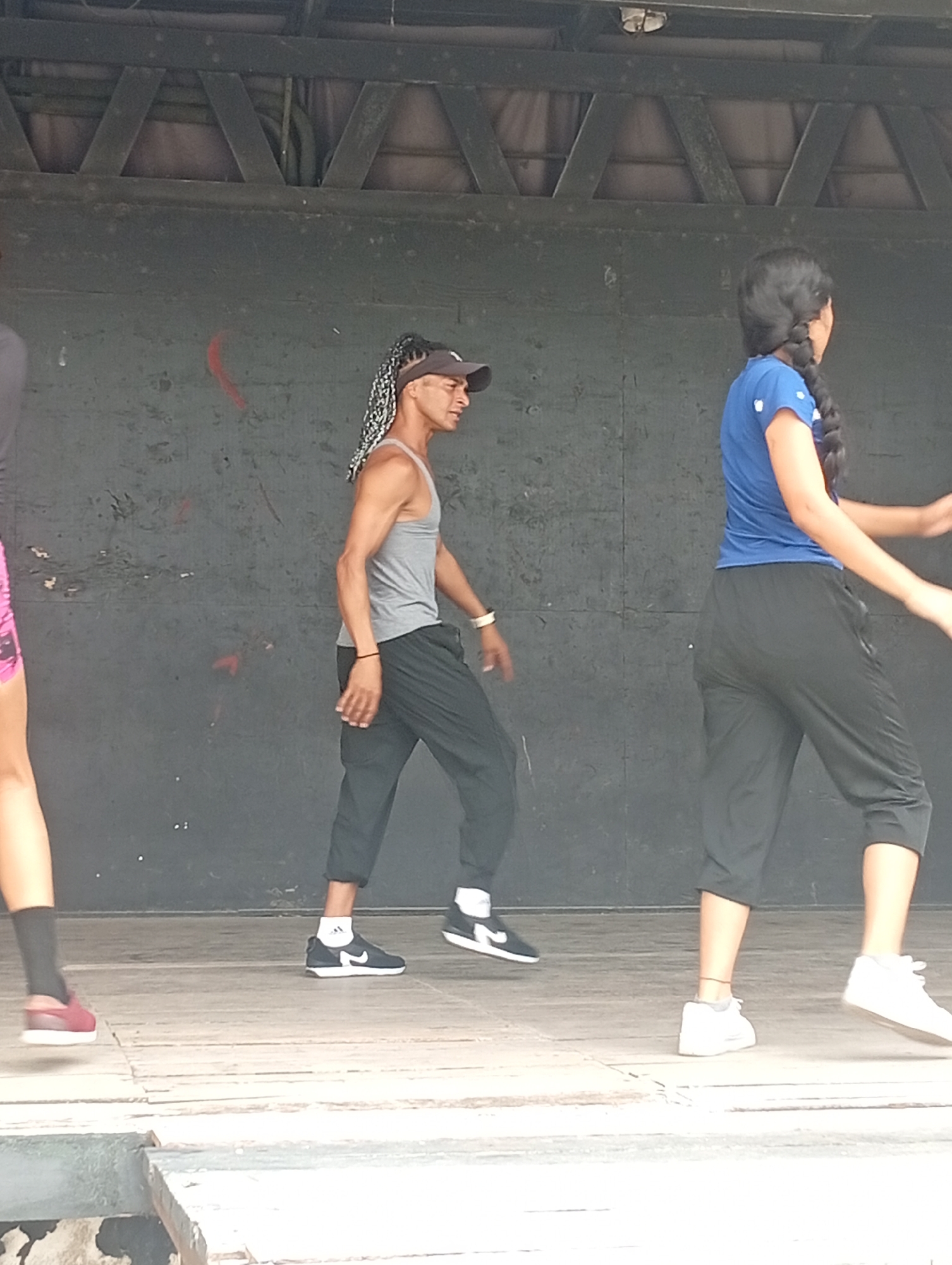 The image depicts three individuals on a wooden stage in an outdoor setting. The backdrop appears to be a dark, possibly painted or weathered wall with a wooden structure above. On the left, a person is partially visible, wearing pink shorts and black socks with maroon sneakers. In the center, a man wearing a grey tank top, black pants, a black visor, and black and white Nike sneakers is walking. He has braids hanging down his back. To the right, a woman with a long braid is wearing a blue shirt and black capri pants, with white sneakers. They are all walking or dancing on the stage. The location may be Playa El Agua, Venezuela, as requested, although there are no landmarks in the image to confirm this.