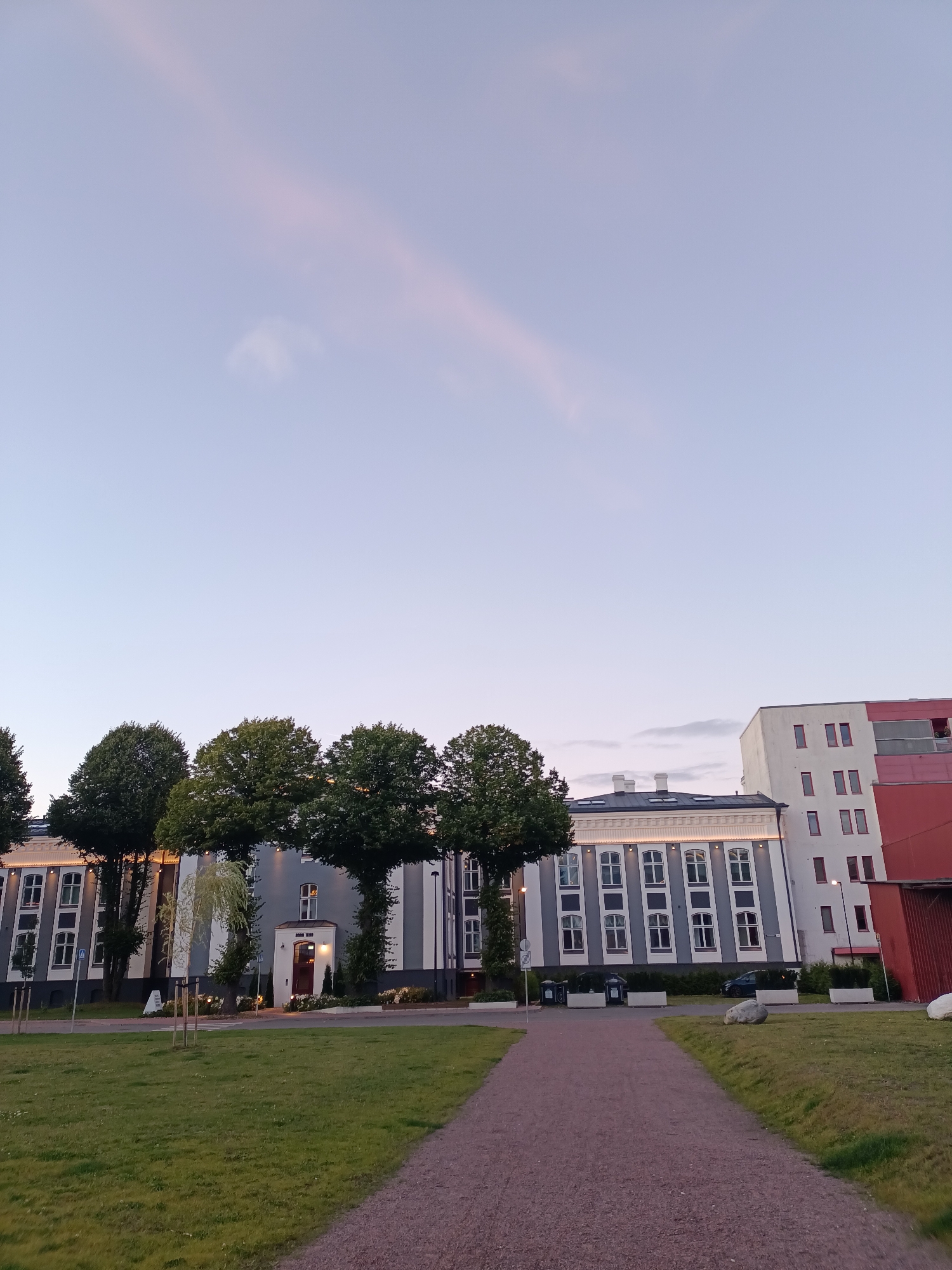 This is a low-angle, wide shot of an urban setting in Tallinn, Estonia, at dusk. The sky is a pale blue with subtle pink clouds. In the foreground, a gravel path leads towards a row of buildings. To the left of the path is a grassy lawn with young trees planted in rows.
The main building in the center is a classical-style structure with a grey facade, white columns, and numerous tall, rectangular windows. A bright light emanates from a recessed entranceway with a dark red door. The building to the right is a modern, multi-story apartment complex with a white and red exterior. A dark red metal-clad structure, possibly a shed or garage, is visible to the far right.
Several mature trees with full, dark green canopies are spaced along the front of the classical building, obscuring parts of its facade. In the background, the sky is clear with a thin streak of pink cloud. The overall scene suggests a quiet evening in a well-maintained urban park or plaza adjacent to residential and historical buildings. There are no people visible in the image.