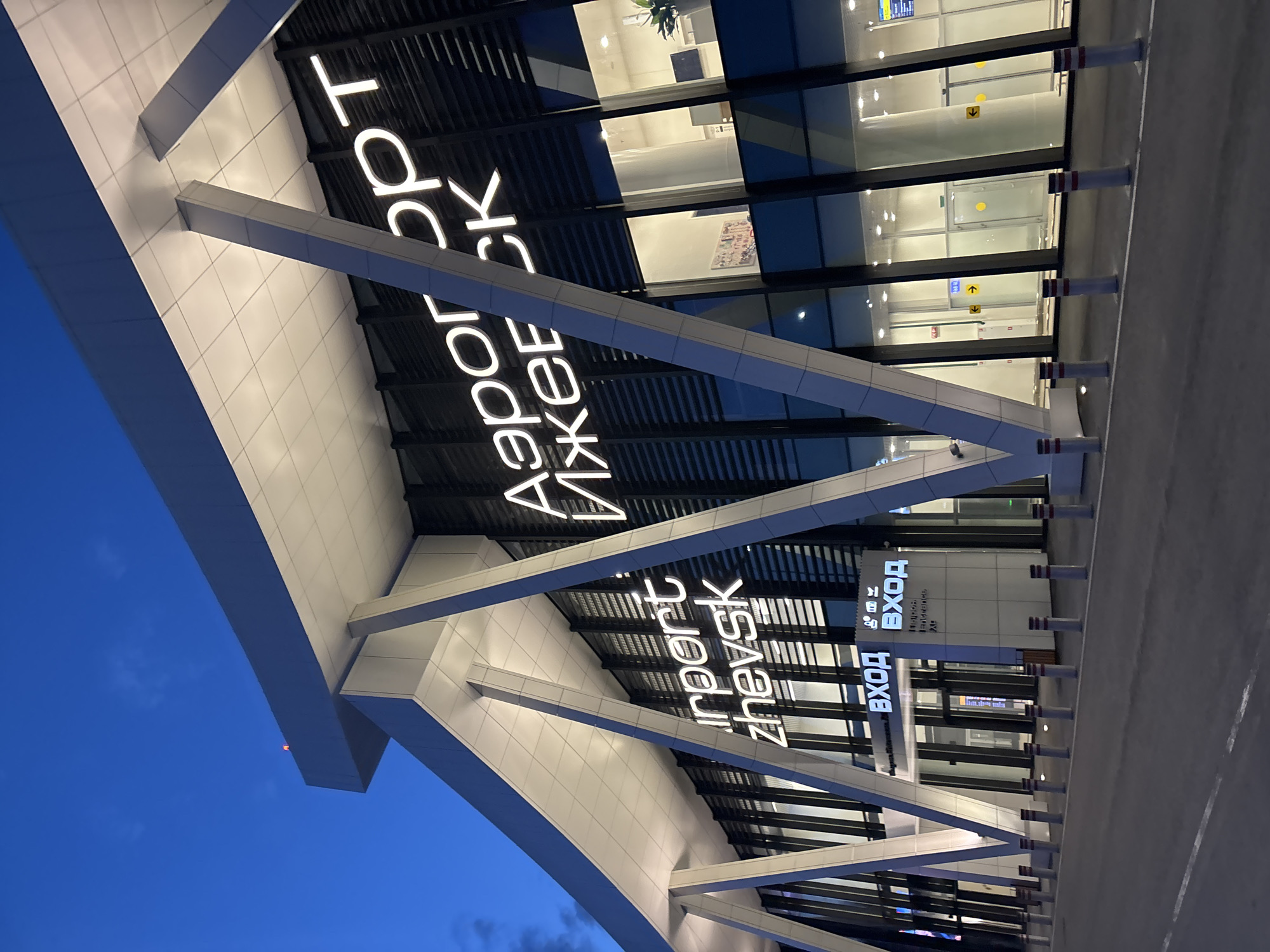 The image shows the entrance to the Izhevsk Airport (Аэропорт Ижевск) in Novo-Martyanovo, Russia. The facade features prominent signage with the airport's name in Russian and English. The building has a modern design with large glass windows and supporting architectural structures. The area in front of the entrance includes bollards for traffic control. The sky is a deep blue, suggesting it is either dusk or dawn.