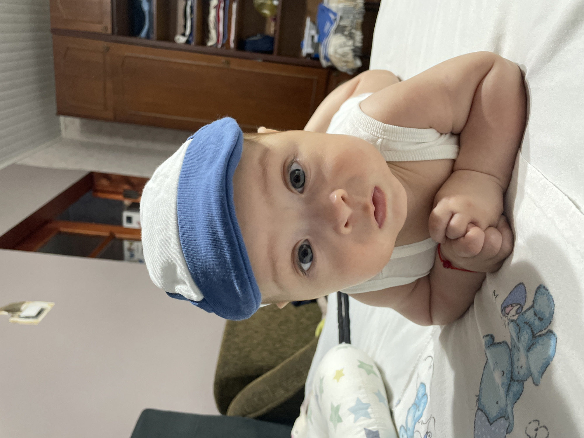 A baby is lying on its stomach on a white surface, wearing a white tank top and a blue and white striped hat. The baby has light skin and blue eyes, and is looking up at the camera with a neutral expression. The background is blurred but appears to be a room with wooden furniture. A red string is tied around the baby's left wrist. The scene is brightly lit, suggesting daytime. The overall impression is one of a relaxed and intimate moment.