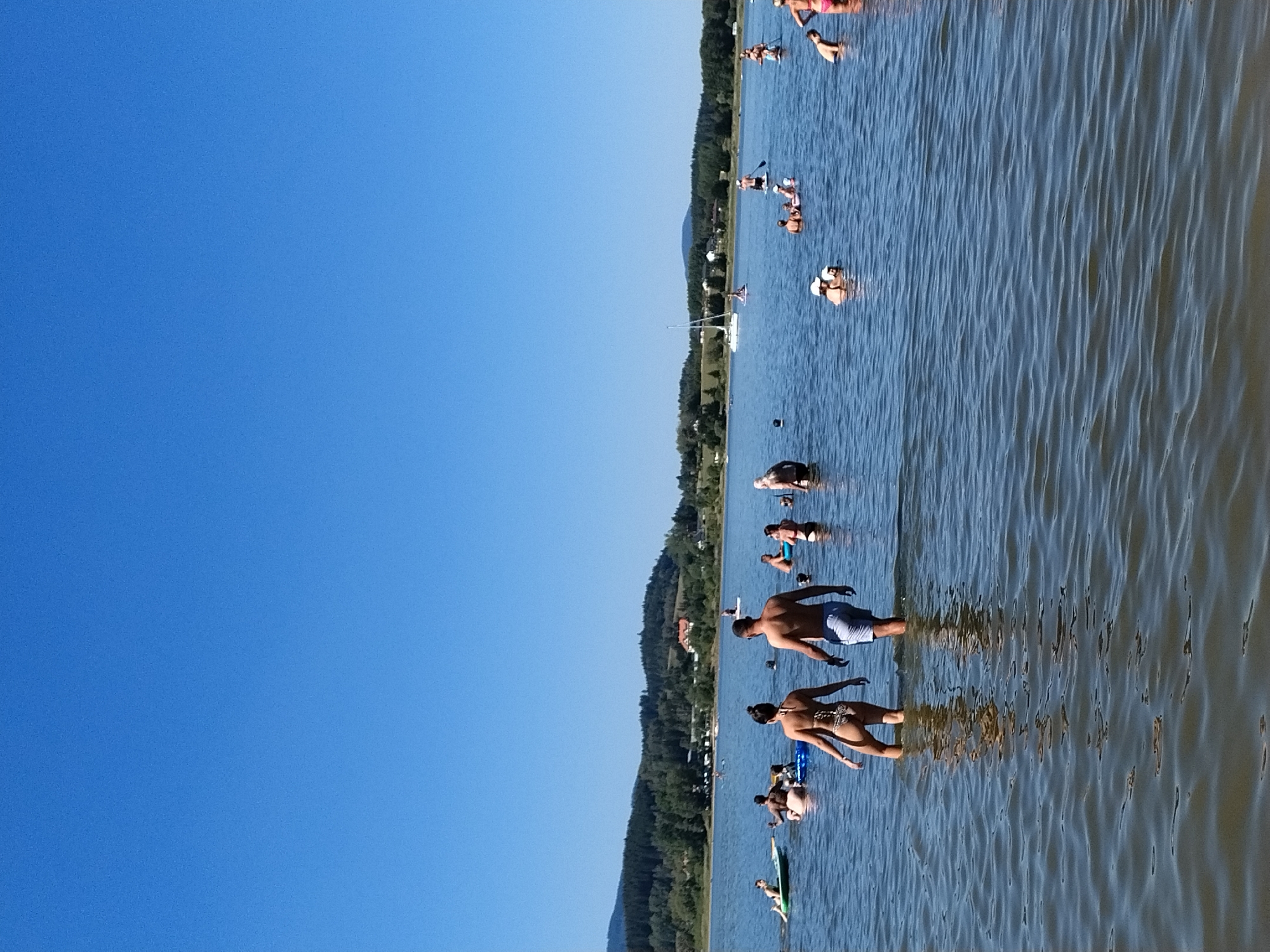 This image captures a sunny day at a lake, likely in or near Radslav, Czechia, given the hilly, tree-covered landscape and a prominent white sailboat in the mid-ground. The water is calm with a few ripples, reflecting the clear blue sky. Several people are enjoying various water activities. In the foreground, a couple walks out of the water, with the woman in a patterned bikini and the man in blue swim trunks. Further out, individuals are seen swimming, relaxing on inflatable mats, and some appear to be paddleboarding. A group of people is clustered together in the middle of the lake, possibly socializing or engaged in a shared activity. The shoreline is lined with trees and some buildings are visible in the distance, suggesting a developed area around the lake. The overall atmosphere is one of recreation and leisure on a warm day.