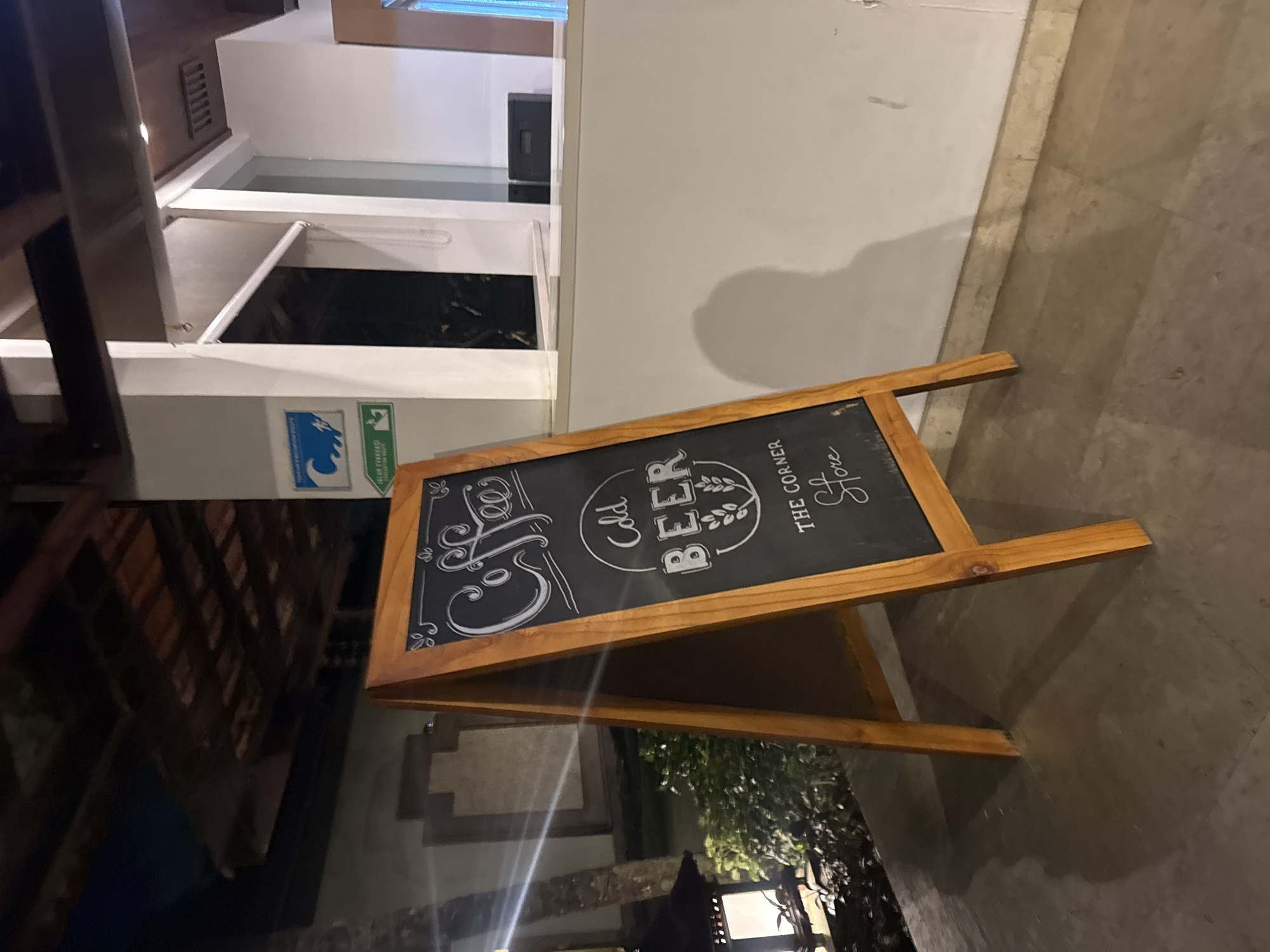 A chalk sign on an A-frame easel stands on a tiled outdoor patio. The sign reads "Coffee," "Cold Beer," and "The Corner Store." Behind the sign, a modern building with white walls and dark wood accents is visible. To the left of the sign, a dimly lit outdoor lamp and lush green foliage are present. Further to the left, a part of a tree trunk is visible. The background shows a section of the building's exterior, including a window and what appears to be a reception desk. The lighting suggests it is nighttime. The location is the exterior of "The Corner Store" in Kuta Selatan, Indonesia.