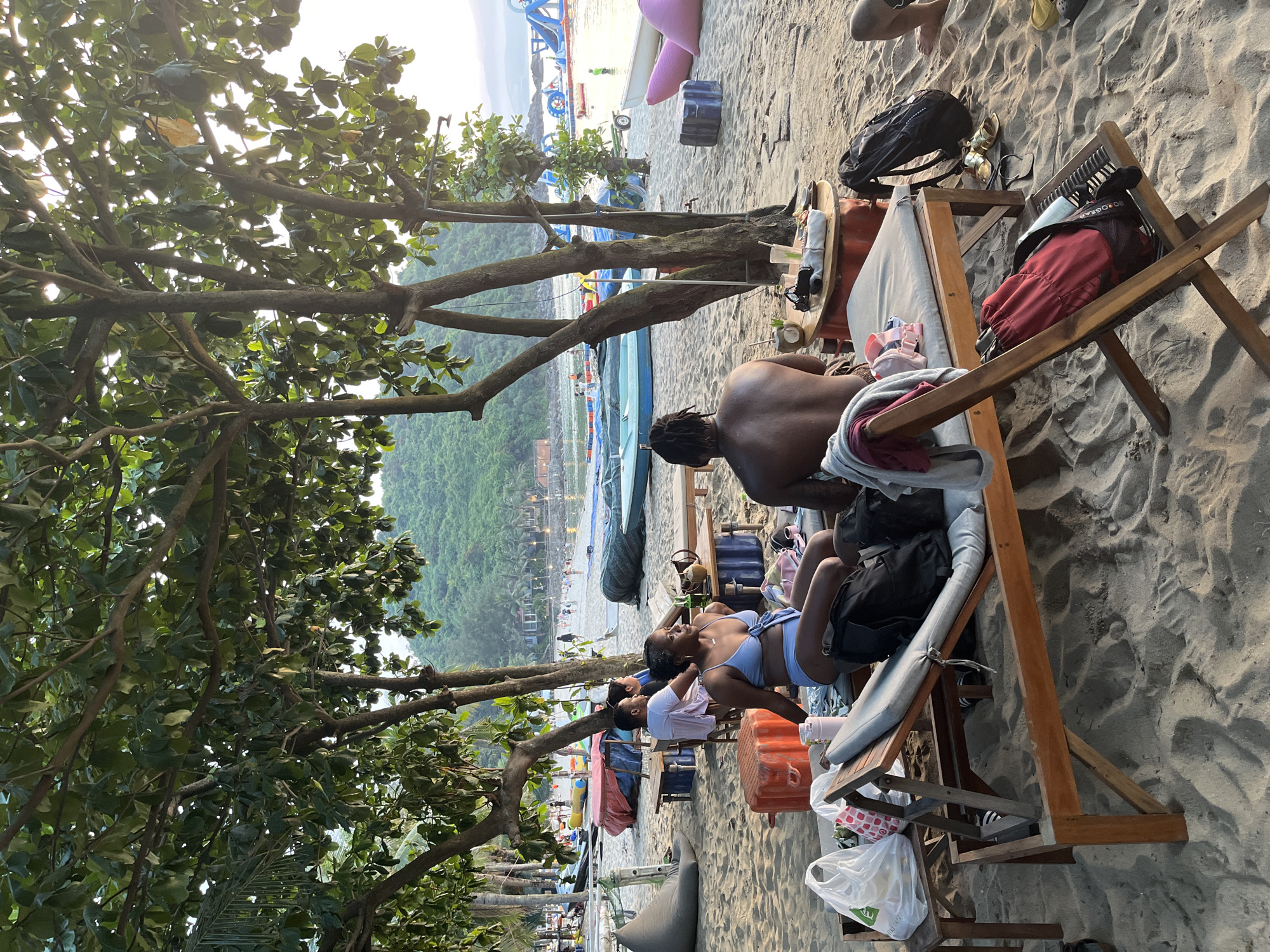 This is a beach scene in Đà Nẵng, Vietnam. A group of people relax on beach loungers under the shade of trees. One man sits shirtless, while a woman in a blue swimsuit reclines on an adjacent lounger. Other people are visible further down the beach. Beach umbrellas, chairs, and various items like bags and bottles are scattered around. In the background, there's a view of the sea, with boats and water equipment visible, along with a green hillside.