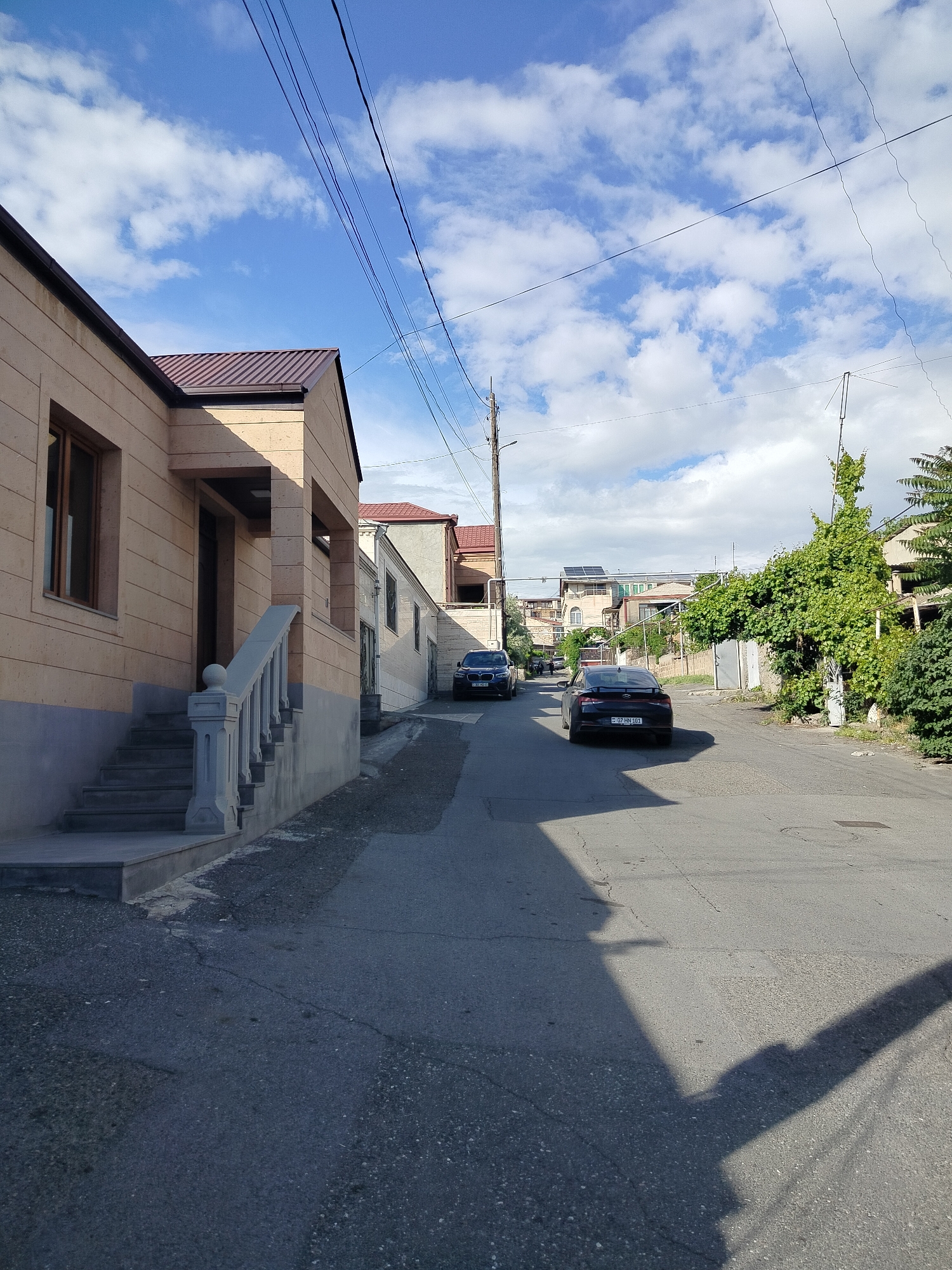 The image depicts a street scene in Yerevan, Armenia. A two-story, light-colored stone building with a red tile roof stands on the left. A set of stairs leads to the entrance, and a balustrade runs alongside. Adjacent to it is a similar building. The street slopes uphill. Two vehicles, a dark-colored sedan and an SUV, are parked on the street. The sedan is closer to the camera. On the right side of the street, there is a fence and overgrown vegetation. The sky is blue with scattered clouds. Power lines are visible overhead. The street appears to be residential.