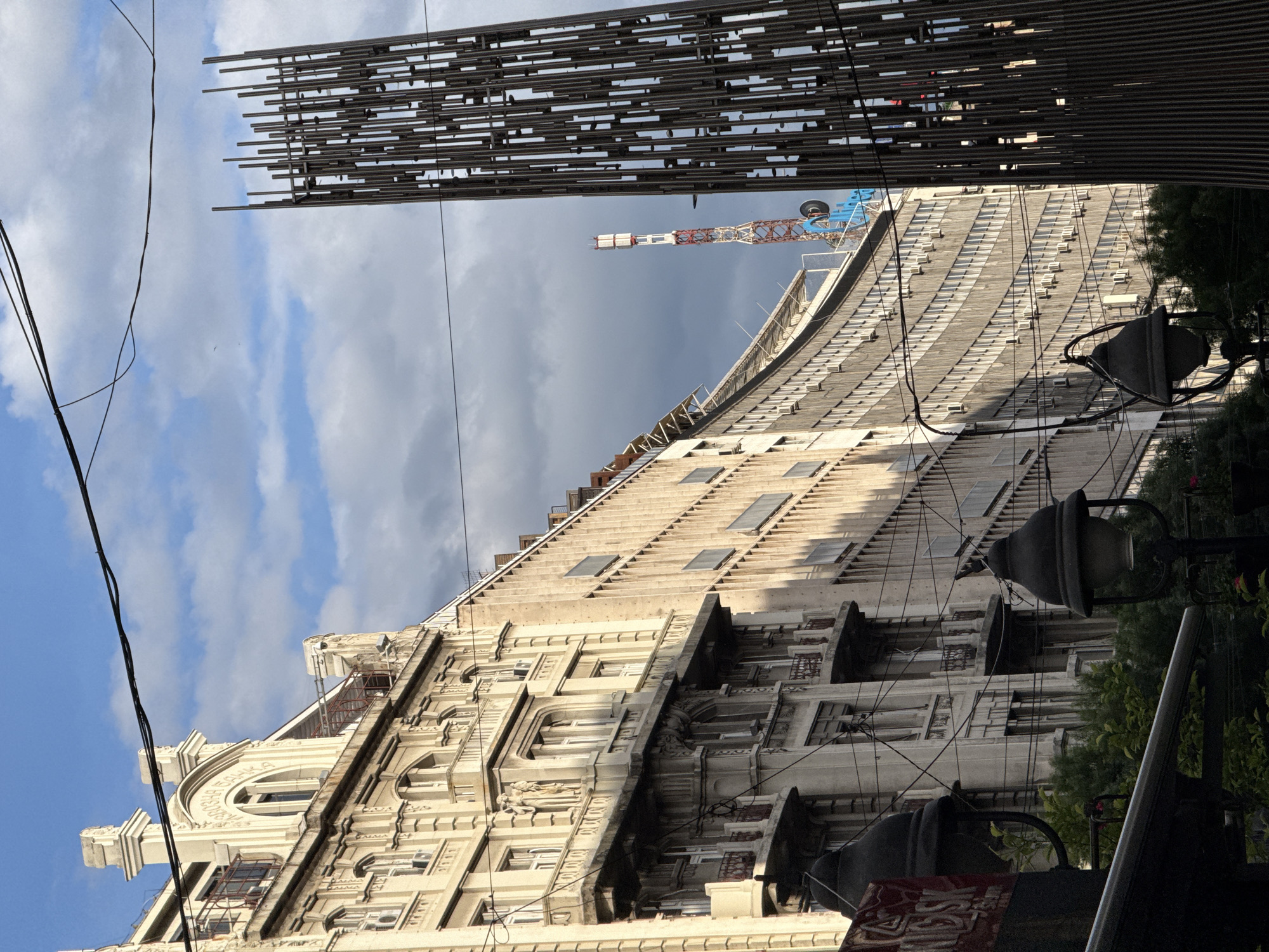 The image shows the facade of a building in Belgrade, Serbia. The building is tall and has intricate architectural details. The top of the building is visible, along with a portion of the roof and a crane. In the foreground, there are street lamps and some greenery. There are electrical wires crossing the frame. The sky is visible, with a mix of blue sky and clouds. Above is what appears to be a metal structure.