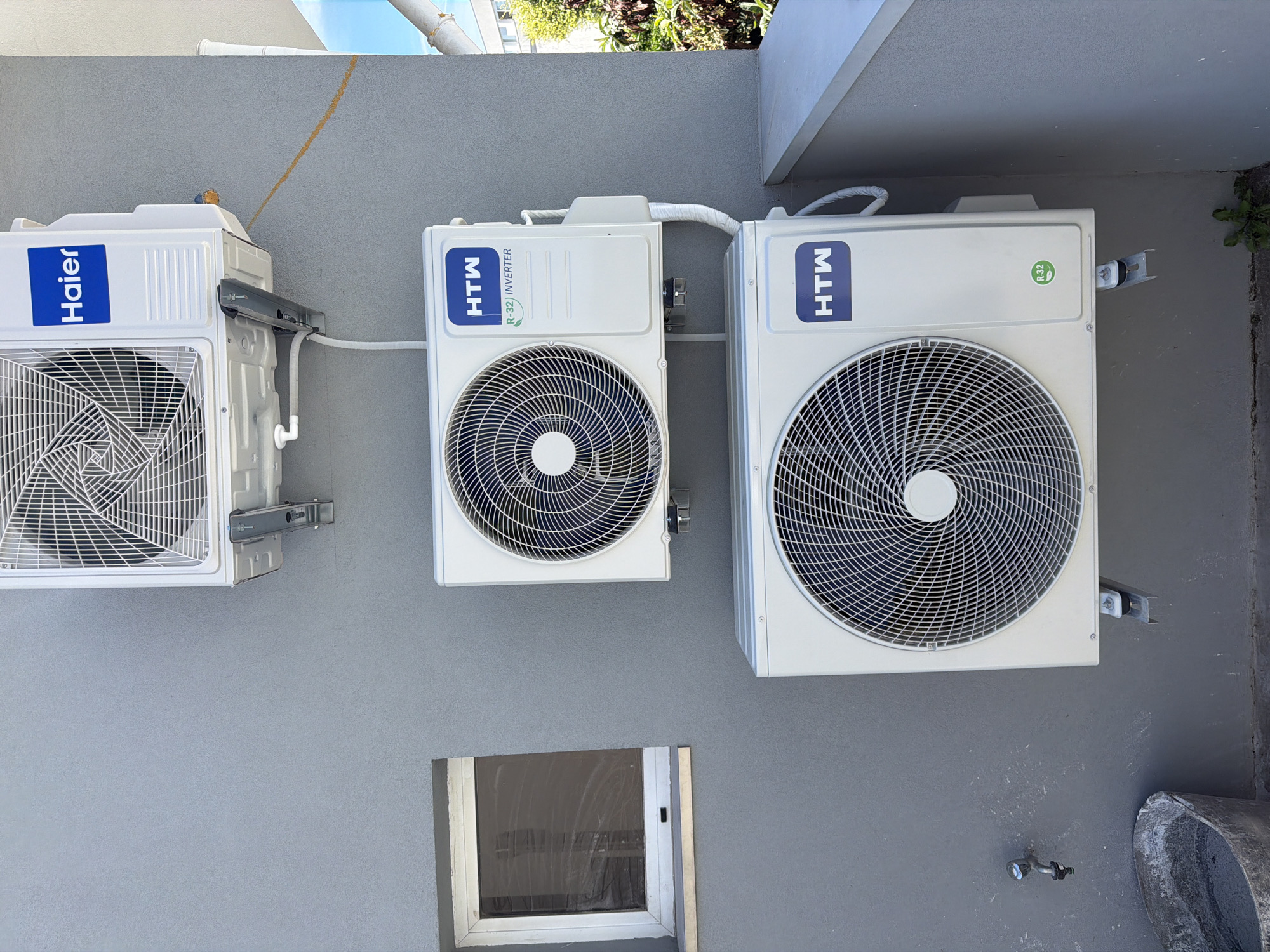 The image shows three outdoor air conditioning units mounted on a grey exterior wall. The topmost unit is a Haier brand, and the two units below it are HTW brand. There is a small, closed window to the left of the units. Below the units, a metal faucet is attached to the wall, and a concrete bucket is placed on the ground. The scene is outdoors, and the wall appears to be part of a building. Based on visual cues in the image, the location is likely in the city of Gondomar, Portugal, where this type of architecture and building materials are common. The image is a static, eye-level shot of the wall and the air conditioning units. There are no people or animals visible. The environment appears to be daytime.