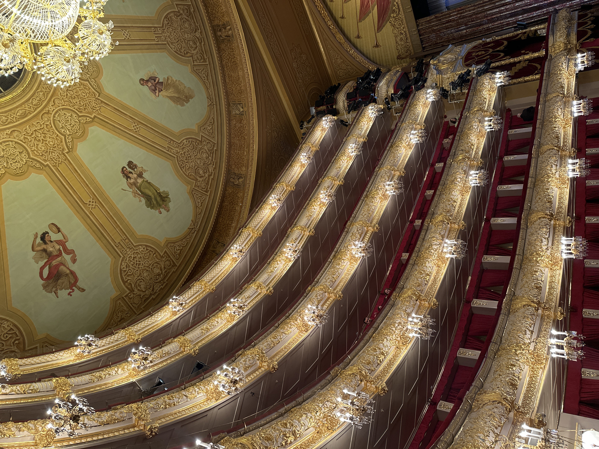 The image captures a dramatic, upward-looking view of the interior of a grand theatre or opera house, showcasing its opulent architecture and intricate decorations. The scene is dominated by multiple tiers of balconies or opera boxes, sweeping in a graceful curve from the left foreground to the right background.
Each tier is adorned with rich, elaborate gold ornamentation featuring highly detailed scrollwork and floral motifs. Along the curved edges of these balconies, numerous small, ornate chandeliers or light fixtures cast a warm glow, illuminating the golden details. Behind the golden railings, the interiors of the boxes are lined with plush red velvet, creating a striking contrast with the gold. Above the upper tiers, professional stage lights and spotlights are visible, mounted on dark structures, suggesting a performance space.
The ceiling, which takes up a significant portion of the upper left, is painted in soft, pastel tones of light green and blue. It is further embellished with gold decorative borders and features several classical-style paintings of figures. One figure in the upper left holds a tambourine and appears to be dancing. Another figure, further up, is depicted in flowing drapery, possibly holding a lyre or similar instrument. A third, less distinct figure is also visible. Large, elaborate crystal chandeliers hang from the ceiling, adding to the grandeur.
The theatre appears largely empty of an audience, with only a few indistinct shapes that might be individuals in the distant upper boxes, suggesting a time before or after a performance, or during a rehearsal. The overall impression is one of historical elegance, luxury, and artistic heritage, consistent with a venue in a city like Moscow. There is no visible text in the image.