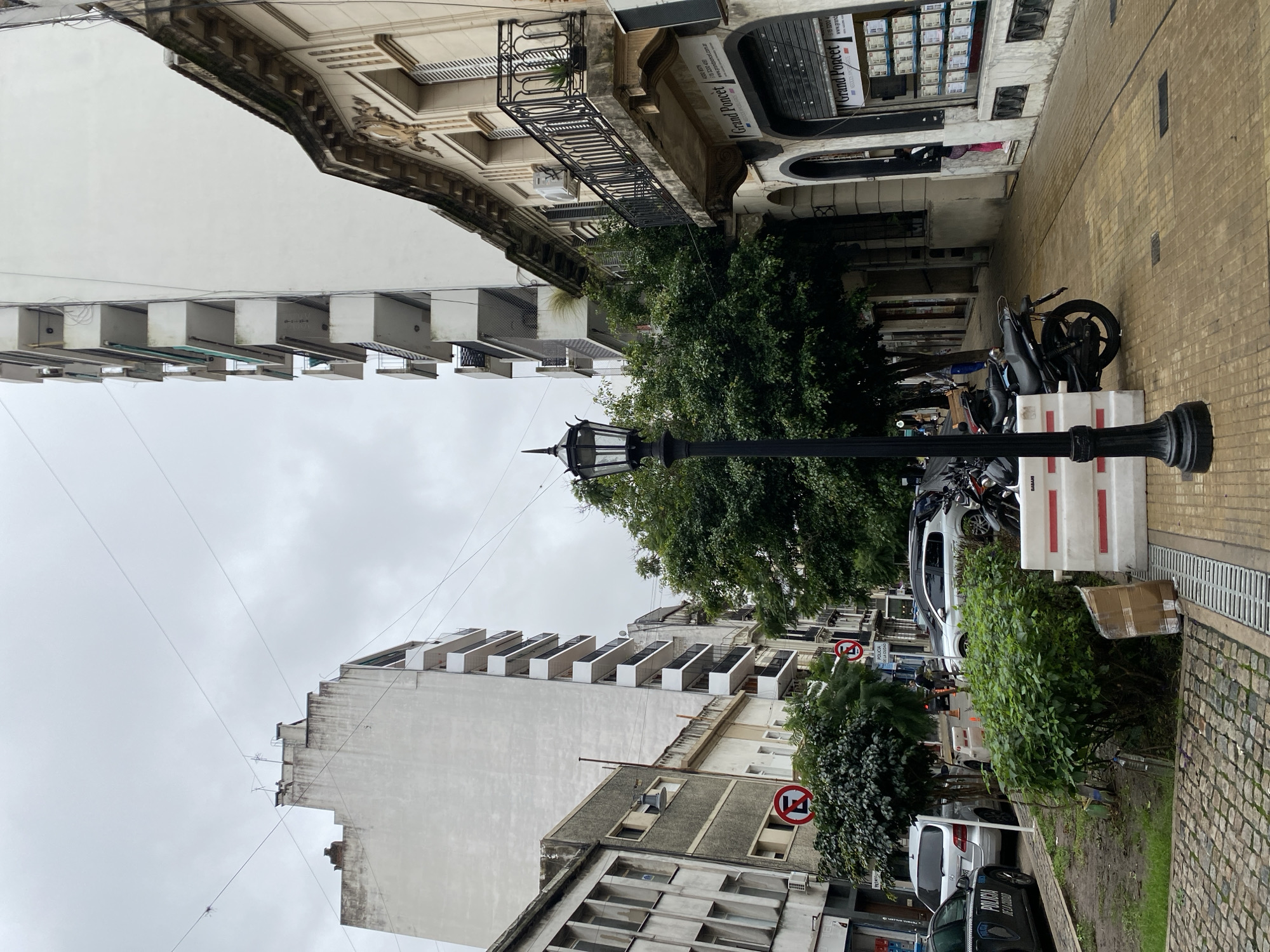 The image depicts a street scene in Buenos Aires, Argentina, with a mixture of architectural styles. Dominating the view are buildings of varying heights. One building has protruding, box-like balconies, while another has classical architectural details, including decorative moldings and a wrought-iron balcony. Street furniture includes a black lamppost with a traditional design. The sidewalk is paved with yellow bricks and includes a drainage grate. On the side of the street is a collection of parked motorcycles and vehicles. One car is labeled "Policia de la Ciudad." Street signs are visible, including "No Parking" signs. A white barrier with red stripes is present. The sky is overcast. The scene is partially obscured by trees and greenery.
