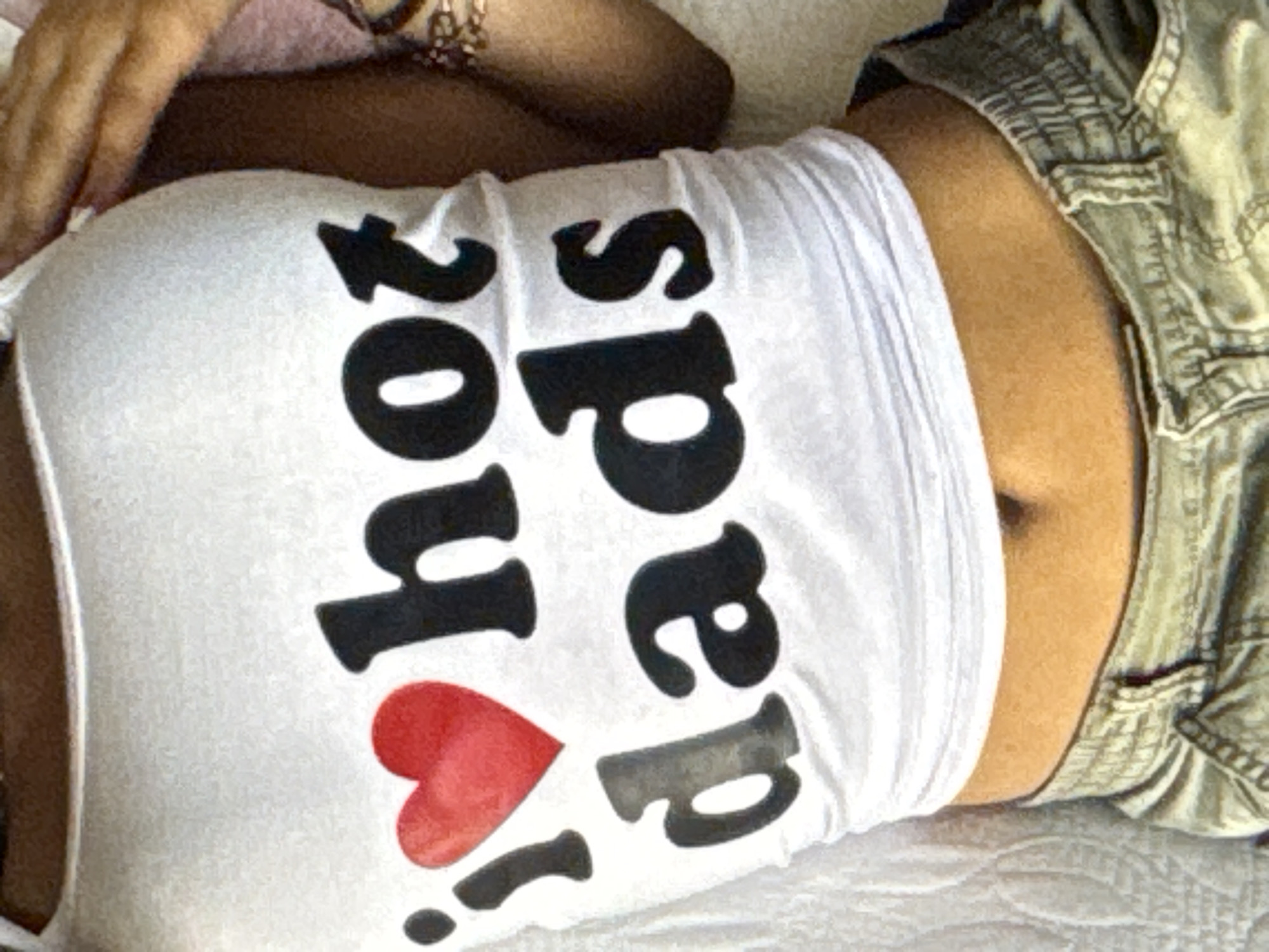 A close-up, overhead shot focuses on the midriff of a person wearing a white crop top with black text and a red heart graphic, and a pair of light-colored cargo shorts. The text on the crop top reads "20 You! Dads." The person's hand, adorned with bracelets, is visible in the upper left corner, resting on a textured pink surface. The background appears to be a bed with a quilted pattern. The lighting suggests it is daytime, and the overall mood is casual. The context is Kuta Selatan, Indonesia.