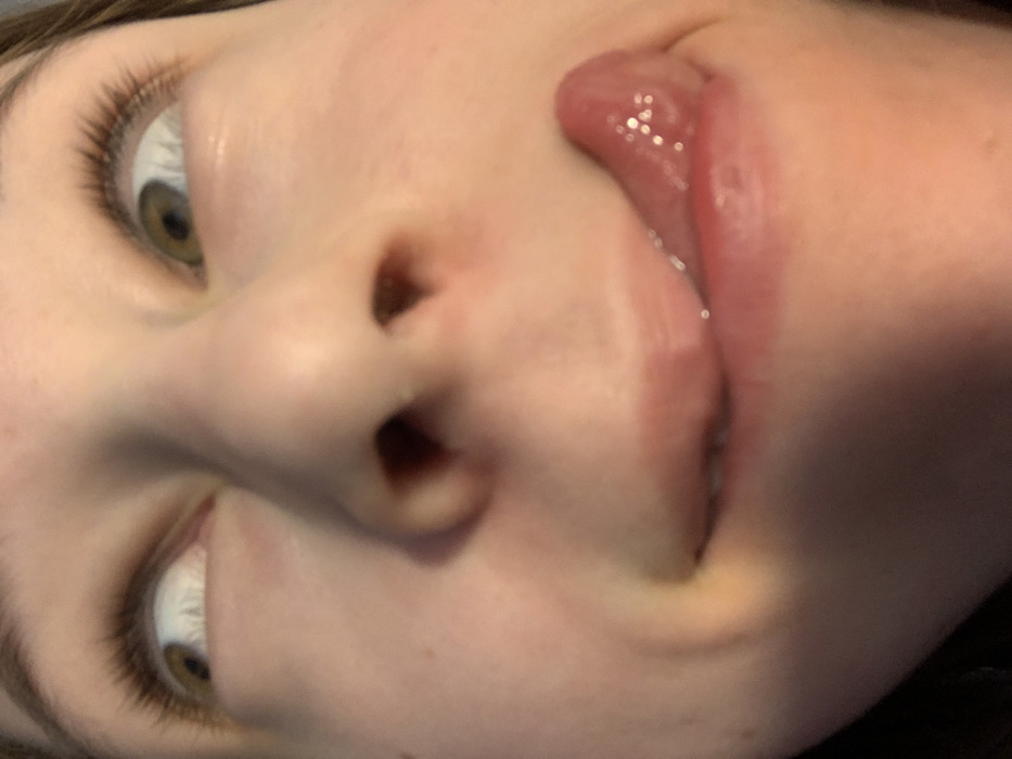 A forensic close-up of an individual's face, set within the city of Vozdovac Urban Municipality, Serbia. The subject, appearing young with fair skin, occupies the majority of the frame. Both eyes are visible, rolled upwards and outwards, displaying light brown or hazel irises and prominent eyelashes. The lower portion of the nose, including the nostrils, is centrally located. The mouth is open, with a moist, reddish tongue extended to the lower right, partially obscuring the lower lip. Superficial skin characteristics include a clear complexion with minor textural variations. Dark hair is visible at the periphery of the upper frame. The depicted action is a deliberate facial expression characterized by upward eye-rolling and lateral tongue protrusion. The immediate environment is undefined due to the extreme close-up perspective and shallow depth of field, offering no discernible background details.
