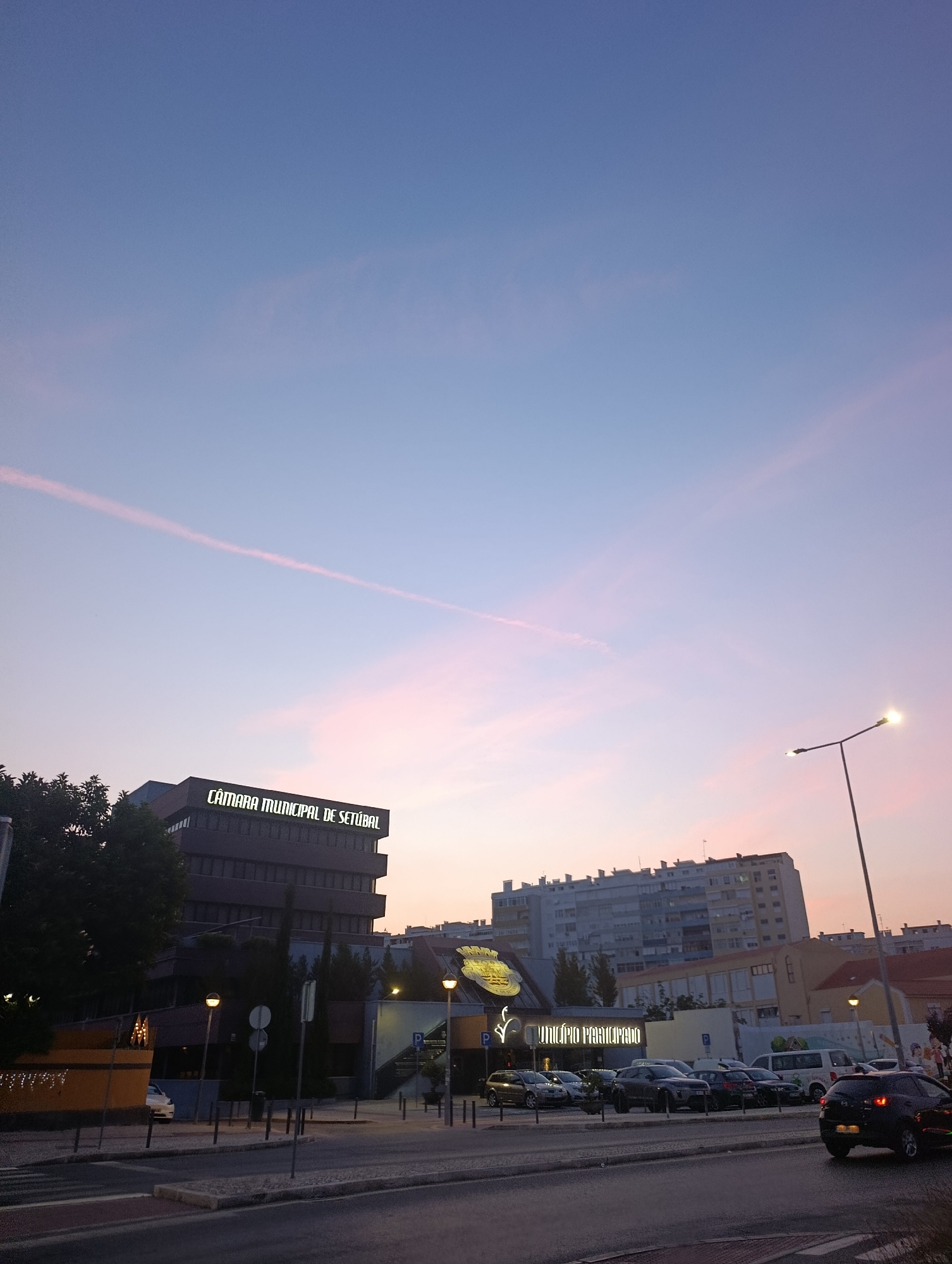 The image depicts a street view in Setúbal, Portugal, at dusk. The sky is a gradient of deep blue at the top transitioning to pink and purple hues closer to the horizon, with a thin contrail visible. On the left, a dark, multi-story building with the illuminated text "CÂMARA MUNICIPAL DE SETÚBAL" across its facade is prominent. Trees are visible in front of this building. Across the street, several cars are parked in front of a building with illuminated signage that reads "MUNICÍPIO PARTICIPADO." Further in the background, a tall, multi-story apartment building stands, and other buildings of varying heights and architectural styles are visible. A street lamp with its light on is positioned on the right side of the image, extending towards the sky. The street in the foreground is paved, with visible crosswalk markings. No people are clearly discernible in the image.