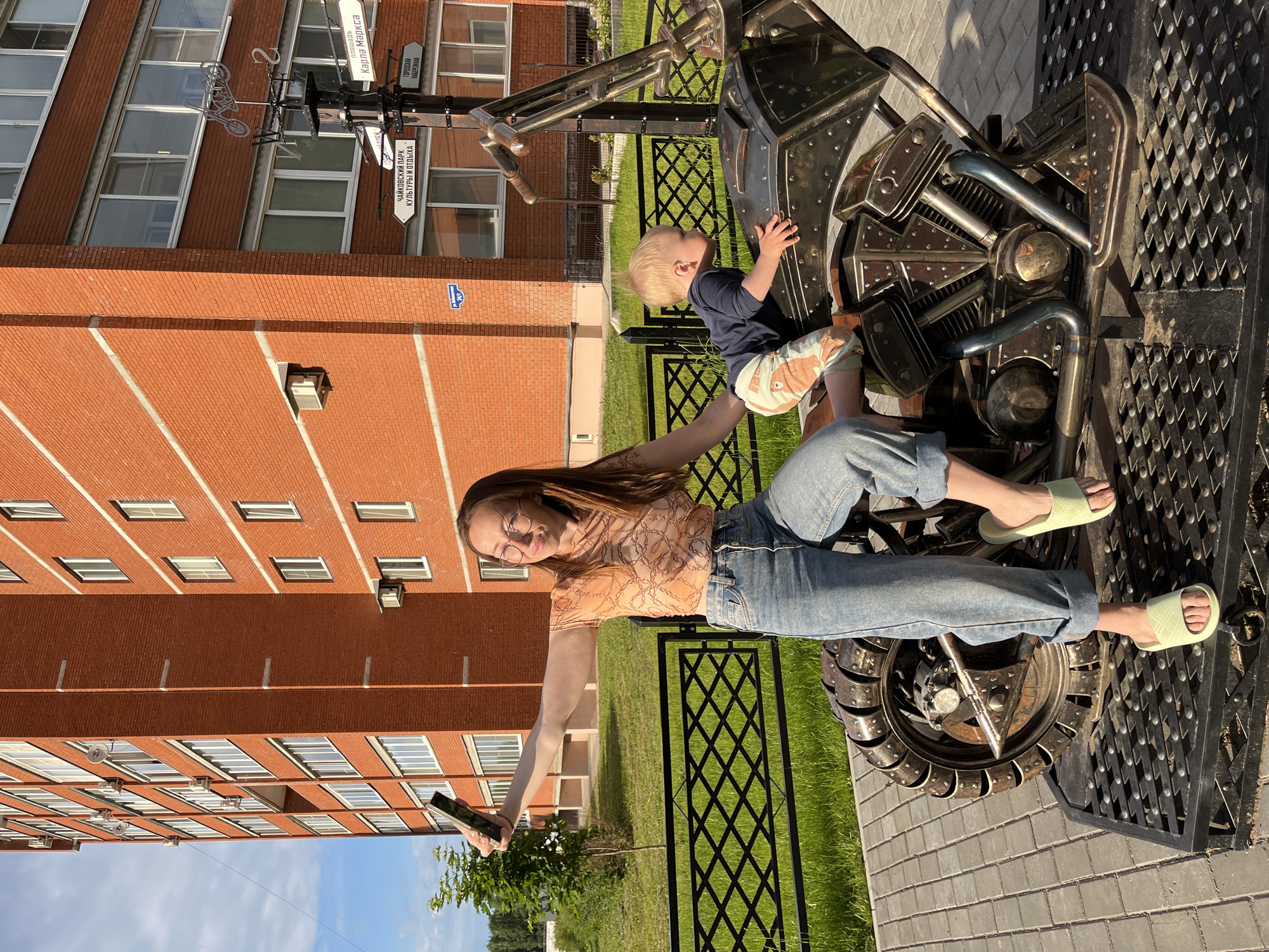 A woman and a young child are interacting with a metal sculpture of a motorcycle in front of a red brick apartment building. The woman, wearing glasses, a patterned t-shirt, and blue jeans, is sitting on the sculpture and taking a selfie with a smartphone. She is wearing light green slippers. The child, with blonde hair, is leaning over the motorcycle. The sculpture is on a metal platform. Behind them, a black metal fence encloses a grassy area. In the background, a multi-story brick apartment building with numerous windows is visible. Street signs in Russian are mounted on a pole near the building. The scene is outdoors, with paved ground and a clear sky, suggesting it is daytime. The location is identified as the city of Chaikovsky, Russia.