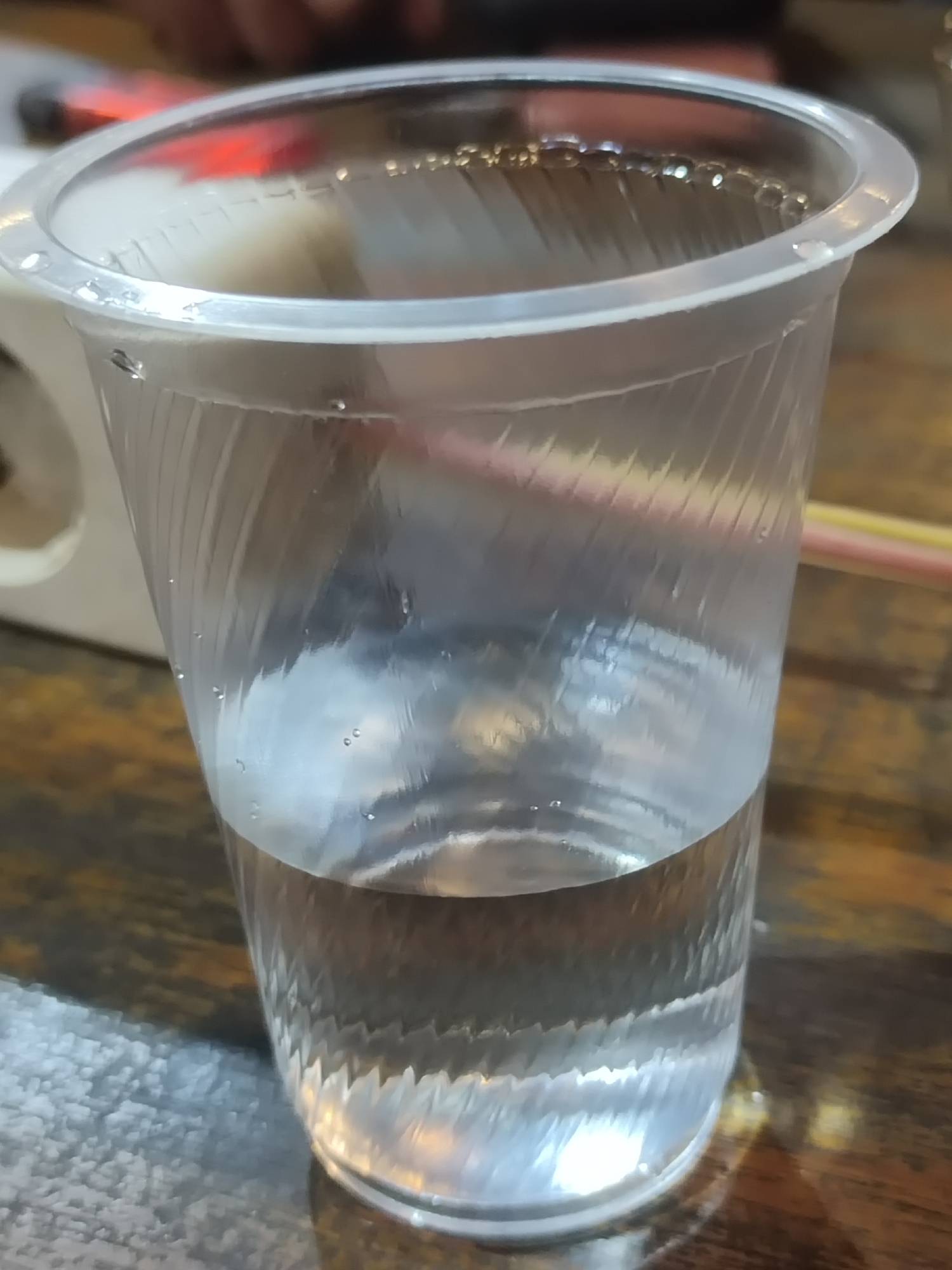 A clear plastic cup filled with a clear liquid sits on a wooden surface. The cup has a ribbed texture and is partially filled. Several small bubbles are visible inside the liquid, and a few droplets of liquid are on the outside rim of the cup. Two straws, one pink and one yellow, are visible behind the cup. The background is blurred, showing a wooden table and a hint of a person's arm. There is no indication of the city of Keudematangkuli, Indonesia in the image.
