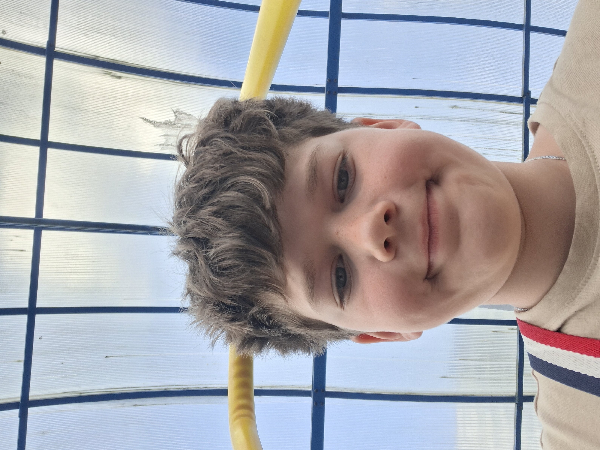 A young boy is depicted from an upward-looking perspective in a close-up shot. He has light skin, messy dark brown hair, and light-colored eyes. Freckles are visible on his nose and cheeks. He has a slight, pleasant smile on his face. He is wearing a light tan t-shirt with a distinctive red, white, and navy blue striped detail on the right shoulder area. A thin silver chain is visible around his neck.
The background shows a translucent, corrugated roof or ceiling structure, likely made of plastic or fiberglass, supported by dark blue metal frames arranged in a grid pattern. Through this translucent material, a bright blue sky is visible, indicating daytime and clear weather. A yellow, curved pipe or railing is partially visible in the upper part of the image, extending diagonally behind the boy's head.
The upward angle of the shot suggests the photograph was taken from below the boy, possibly while he was lying down and looking up, or the camera was pointed upwards from a low vantage point. The setting appears to be an outdoor or semi-enclosed recreational area, such as a covered pool deck or a similar structure in a public park, consistent with the context of Saint Petersburg, Russia. There is no visible text in the image.