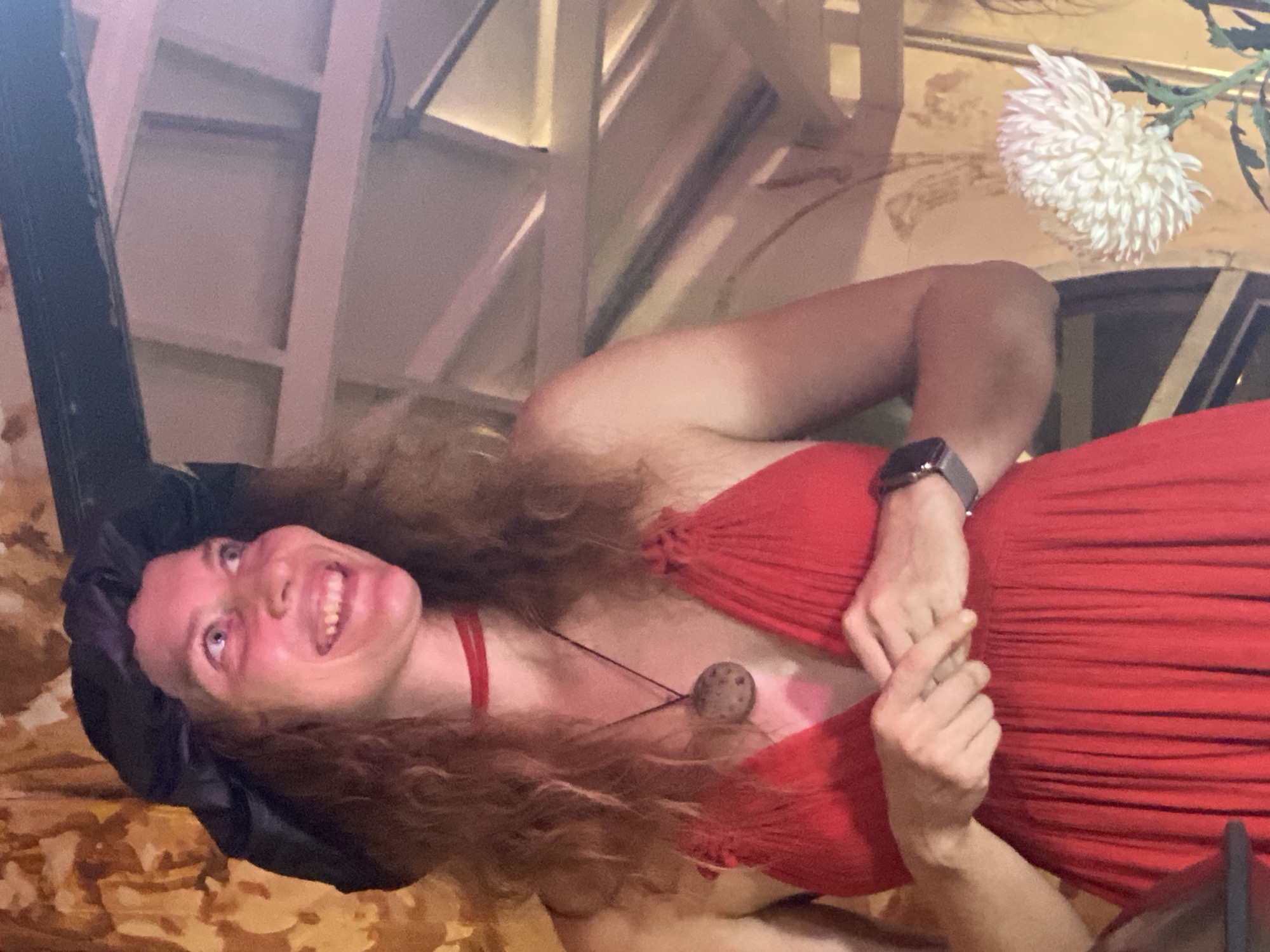 A medium shot captures a woman standing indoors, positioned slightly off-center to the left, in front of a patterned wall and a white flower. She is wearing a red dress with a V-neck and thin straps, a black head covering, and a thin red choker necklace with a circular pendant. Her curly, auburn hair is visible around her face and shoulders. The woman is smiling broadly and looking up and to her right, with her hands clasped together in front of her chest. A smartwatch is visible on her left wrist. The environment includes a visible ceiling structure and ambient lighting that casts a reddish hue on the scene. The white flower, likely a chrysanthemum, is in the lower right portion of the frame.