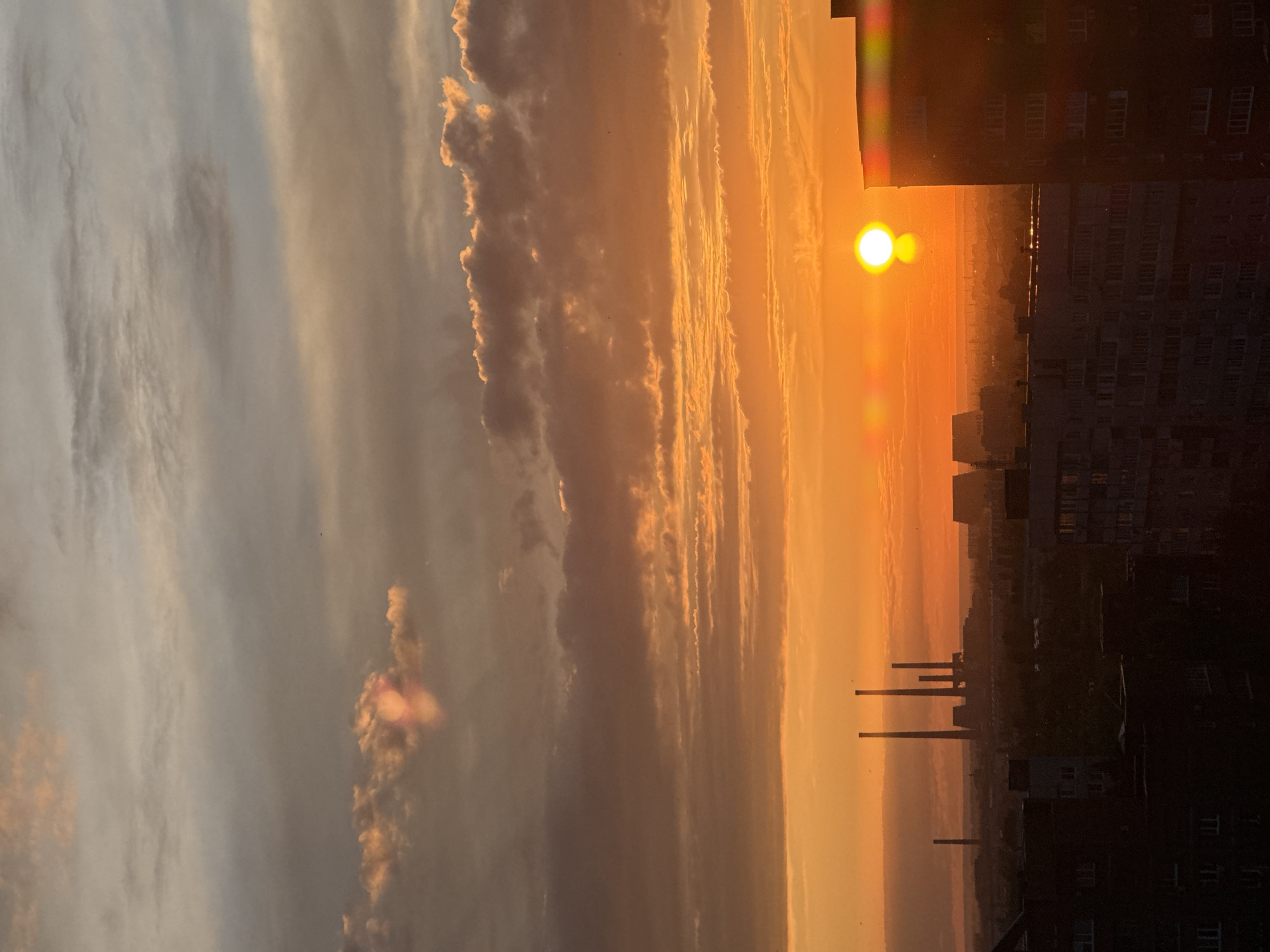 The image captures a panoramic view of the city of Unknown, Unknown, during either sunrise or sunset. The sky is dominated by intense orange and yellow hues, particularly in the lower half, transitioning to warmer grey and light blue tones in the upper regions. A bright, circular, yellow-white sun disk is visible low on the horizon, slightly right of the frame's vertical center. Distinct lens flares emanate from the sun, appearing as a vertical streak above it and horizontal streaks. Numerous clouds are present, with those closer to the horizon exhibiting warm orange and red illumination, while higher clouds appear as darker grey masses. Scattered small, dark specks, consistent with birds, are observable across the mid-sky.
In the midground, several tall, slender industrial chimneys or smokestacks are silhouetted against the bright sky on the left side of the frame. The distant cityscape forms a hazy, dark band along the horizon line. The foreground is dominated by the dark silhouettes of multi-story urban buildings. A large, rectangular building on the right side of the foreground is heavily shadowed, partially obscuring the sun, with visible windows and interacting with lens flare effects along its right edge. No individuals are discernible within the frame.