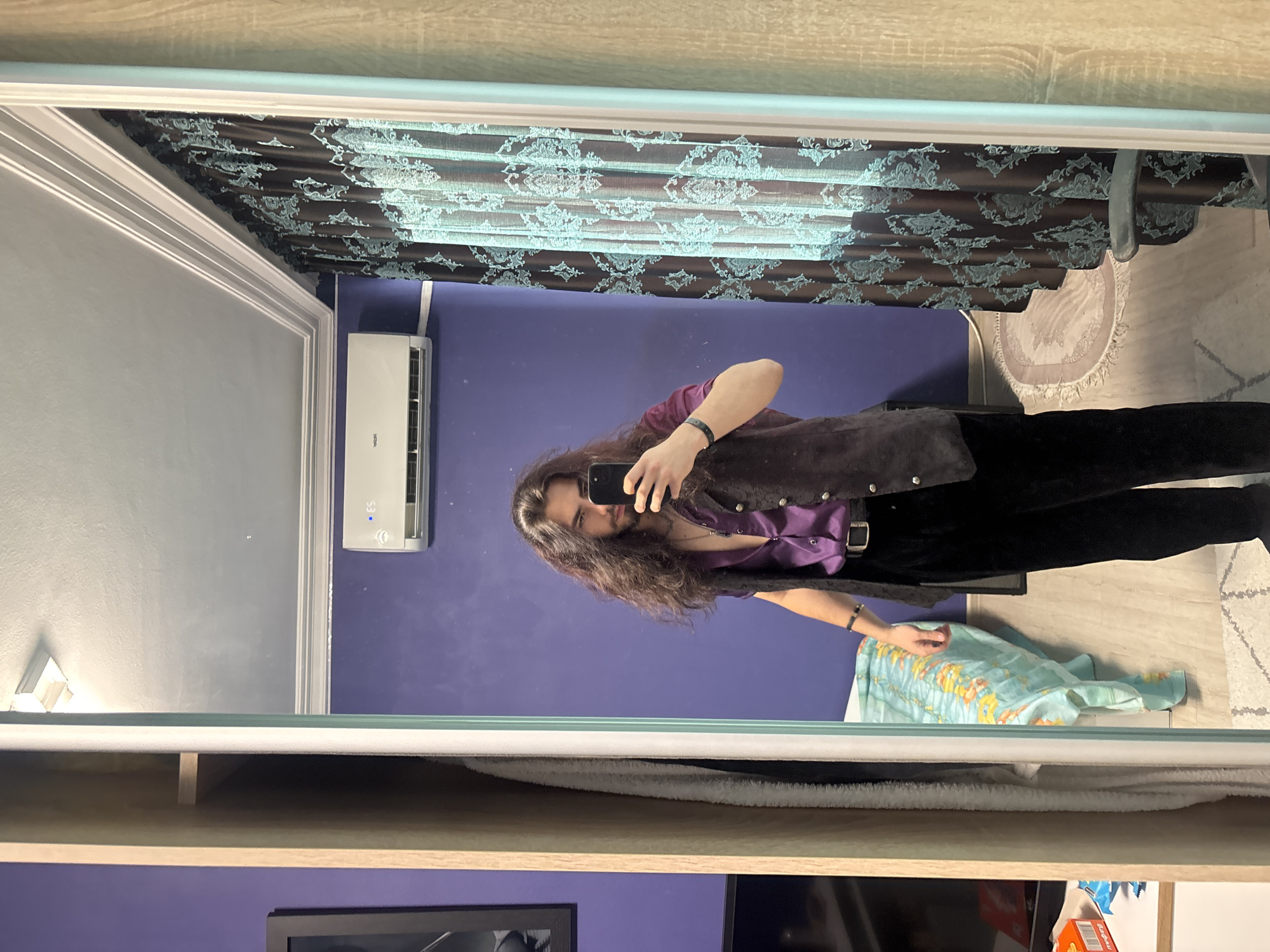 A young man with long, dark, wavy hair and a beard stands in front of a large mirror, taking a selfie with a smartphone. He is wearing a purple satin button-up shirt, a dark vest, black trousers with a silver buckle belt, and a necklace with a cross pendant. He has a bracelet on his left wrist. The mirror reflects the room behind him, which includes a blue wall, an air conditioning unit, patterned curtains, a bed with a light blue and yellow patterned comforter, and a small rug on the floor. To the left of the mirror, a portion of a closet with shelves is visible, containing a white towel or blanket and some boxes on a shelf. The flooring is light wood-colored laminate.