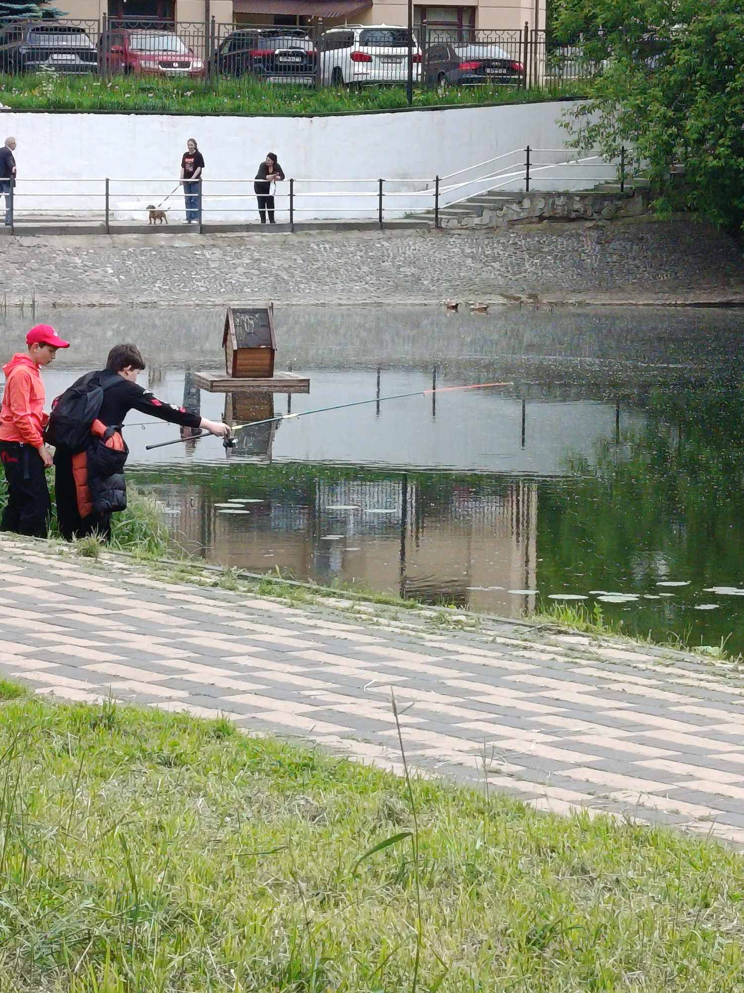 This image captures a daytime scene along a body of water in Moscow, Russia, possibly a pond or canal. In the foreground, two boys are engaged in fishing. One boy, wearing a red cap and an orange top, stands beside another boy dressed in black with a backpack, who is actively holding a fishing rod extended over the water. A small, wooden house-like structure is visible on a platform in the water further out.
The immediate foreground features a patch of green grass and a checkered paved path running parallel to the water's edge. The water itself reflects the surrounding environment, including distant buildings and trees.
On a raised embankment in the background, several people are present. One person is seen walking a small brown dog on a leash, while another leans against a white railing. A third individual is visible further to the left. Behind them, a fence separates the public area from parked cars and residential-style buildings. Green trees and foliage are also prominent on the right side of the background. The lighting is soft and diffused, suggesting an overcast day. No discernible text is visible in the image.