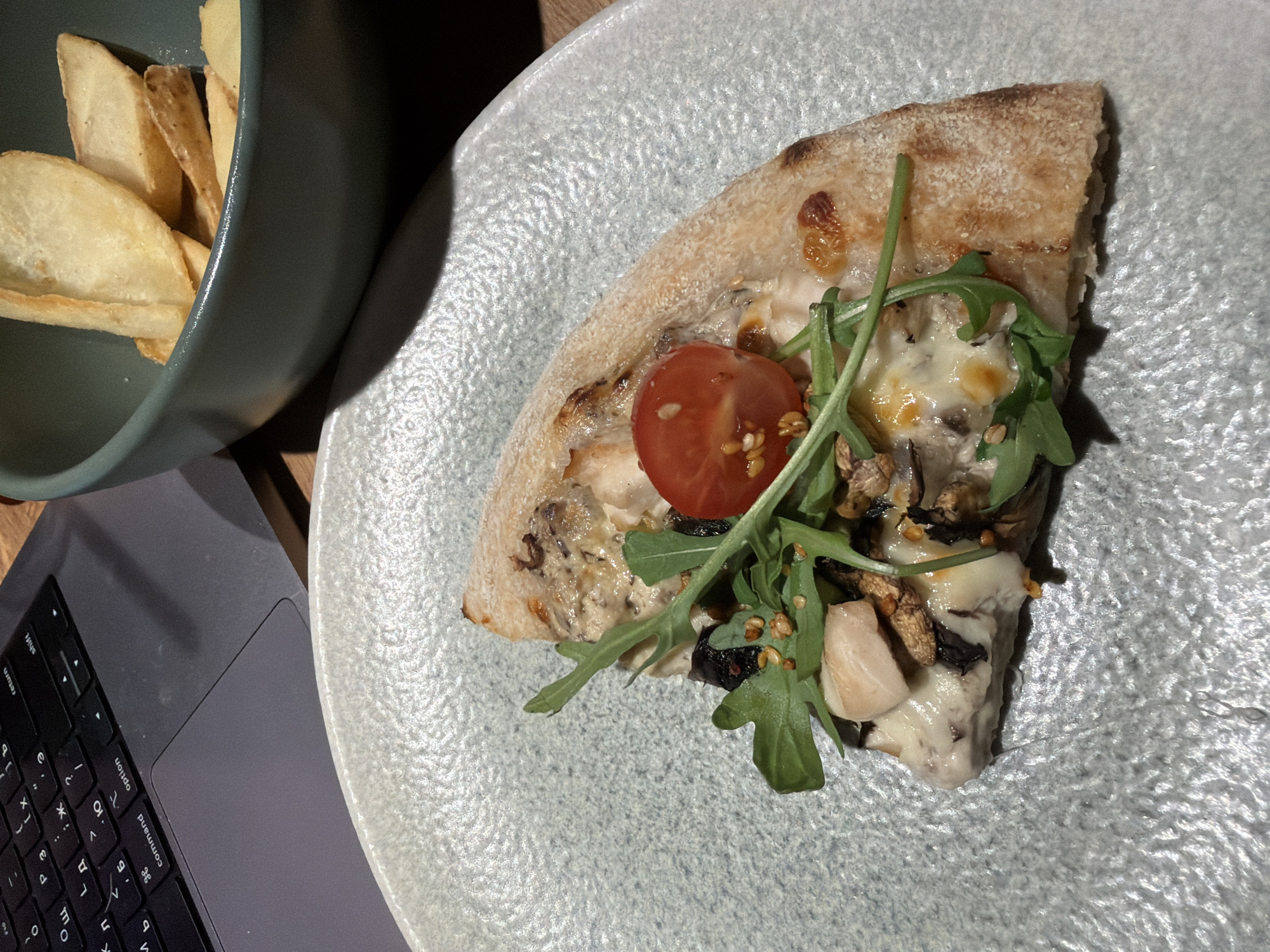 The image presents an overhead view of a meal set next to a laptop, likely in an indoor setting such as a cafe or home in Chaikovsky, Russia.
The primary subject is a single slice of pizza, centrally placed on a round, light grey plate with a distinct textured surface. The pizza slice has a thick, well-baked crust and is generously topped with melted cheese, fresh green arugula leaves, a vibrant halved cherry tomato, and pieces of what appear to be chicken or mushrooms, along with small, dark granular seasonings.
To the upper left, a dark teal or green bowl contains several golden-brown potato wedges. Adjacent to the food on the lower left, a grey laptop, consistent in appearance with a MacBook, is open. Its keyboard is partially visible, displaying both Latin characters like "return," "option," and "command," as well as Cyrillic letters, strongly indicating a Russian-speaking context.
The scene suggests an individual enjoying a meal while engaged with a computer, possibly working or browsing. The lighting is warm and appears to be artificial, casting soft shadows that add depth to the composition. No people are visible in the frame.