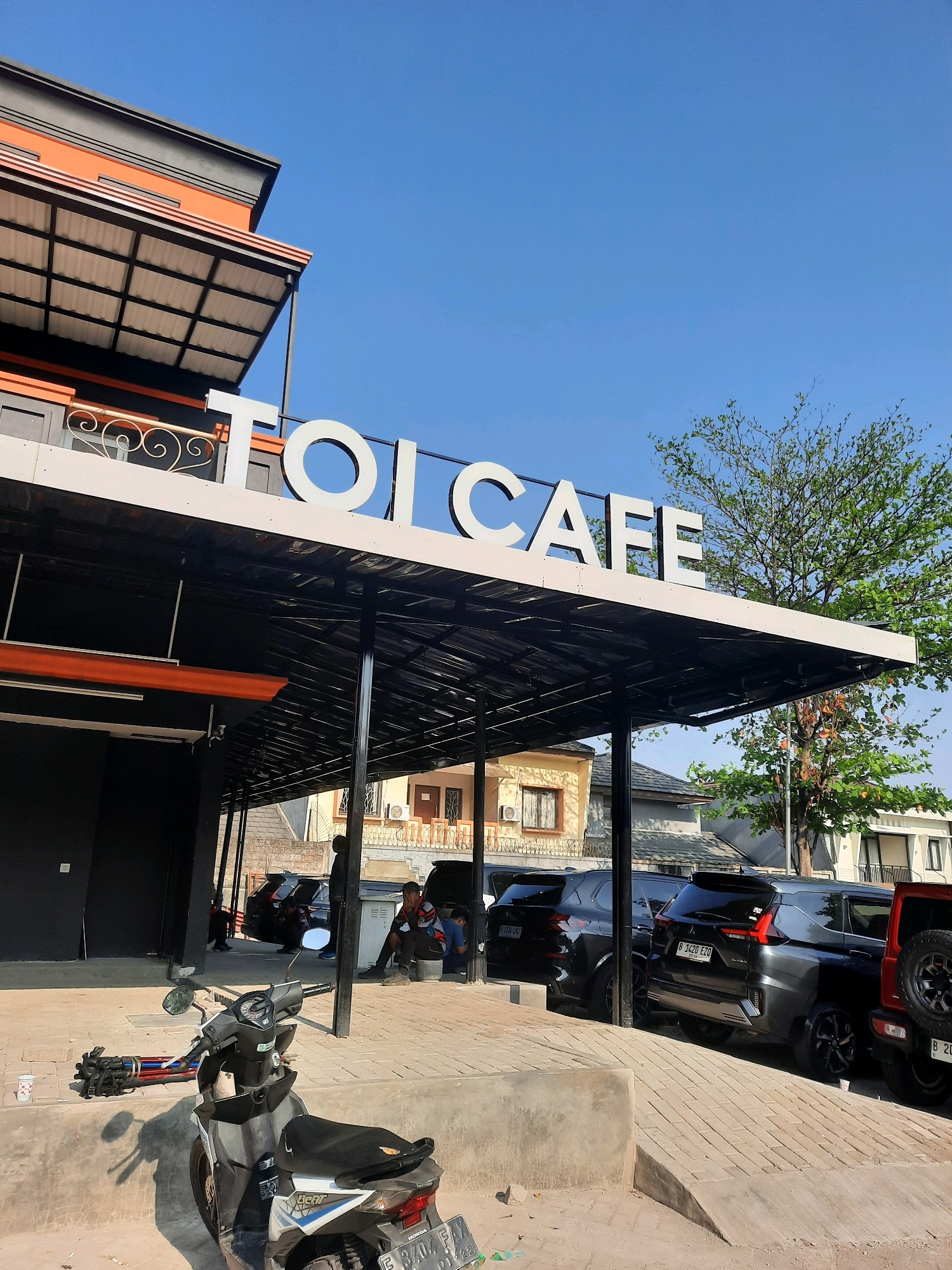 The image depicts the exterior of "TOL CAFE" in Babakan Madang, Indonesia, under a clear blue sky. The cafe features a modern architectural design with a prominent white sign spelling out "TOL CAFE" in large, bold letters. An orange and grey overhang, supported by black pillars, provides shade over a seating area or walkway. Parked vehicles, including several SUVs and a motorcycle, are visible in front of and around the cafe. Two individuals are seated under the overhang, appearing to be customers or staff. The motorcycle in the foreground is a white and black Honda Beat. The overall scene suggests a commercial establishment open for business on a sunny day.