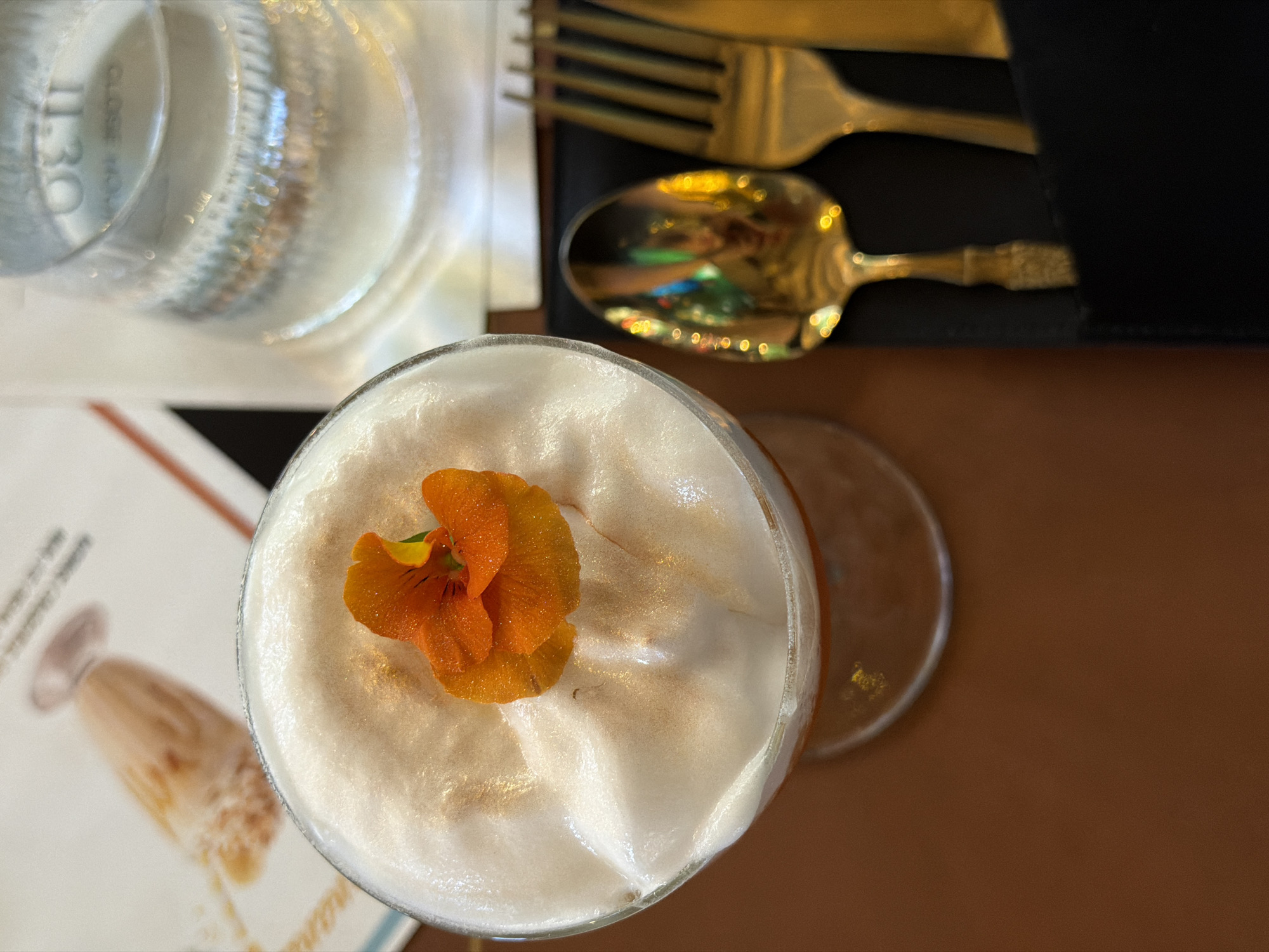 This overhead shot captures a table setting, likely at a restaurant or bar. The central focus is a drink in a clear glass, topped with a thick layer of creamy white foam. An vibrant orange edible flower, possibly a nasturtium, adorns the foam, which also appears to have subtle golden, shimmering specks.
To the upper left, a clear drinking glass, likely containing water, reflects light. Faint text and numbers, including "11:30" and partially "CLOSE HO," are visible on its surface, either embossed or reflected from nearby objects. In the upper right corner, golden-colored cutlery, consisting of a fork and a spoon, rests on a black surface, possibly a napkin or a cutlery holder. The spoon's reflective surface shows a colorful, blurred image, suggesting reflections of the room's interior.
To the bottom left, a white menu or paper is visible, featuring blurred text and an illustration of another drink in a glass. The table surface beneath all these items is a plain, warm brown hue. The overall scene suggests an elegant dining or cocktail experience. The provided context indicates the location is Sofia, Bulgaria.