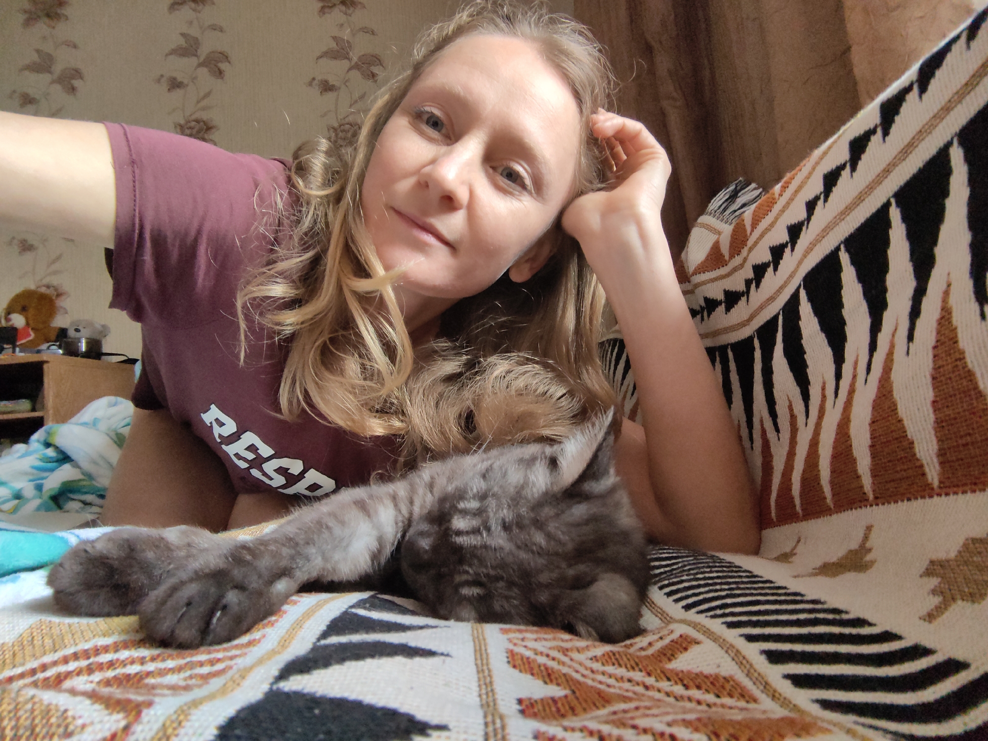 A woman with blonde curly hair is lying down on a patterned blanket, holding a kitten. The woman is wearing a maroon t-shirt with the word "RESP" visible on it. The kitten is gray and appears to be sleeping on the woman's chest. The background shows a floral wallpaper and part of a curtain. The scene suggests a domestic setting, likely a home. The location of Зуйское сельское поселение, Ukraine is not discernible from the image itself.