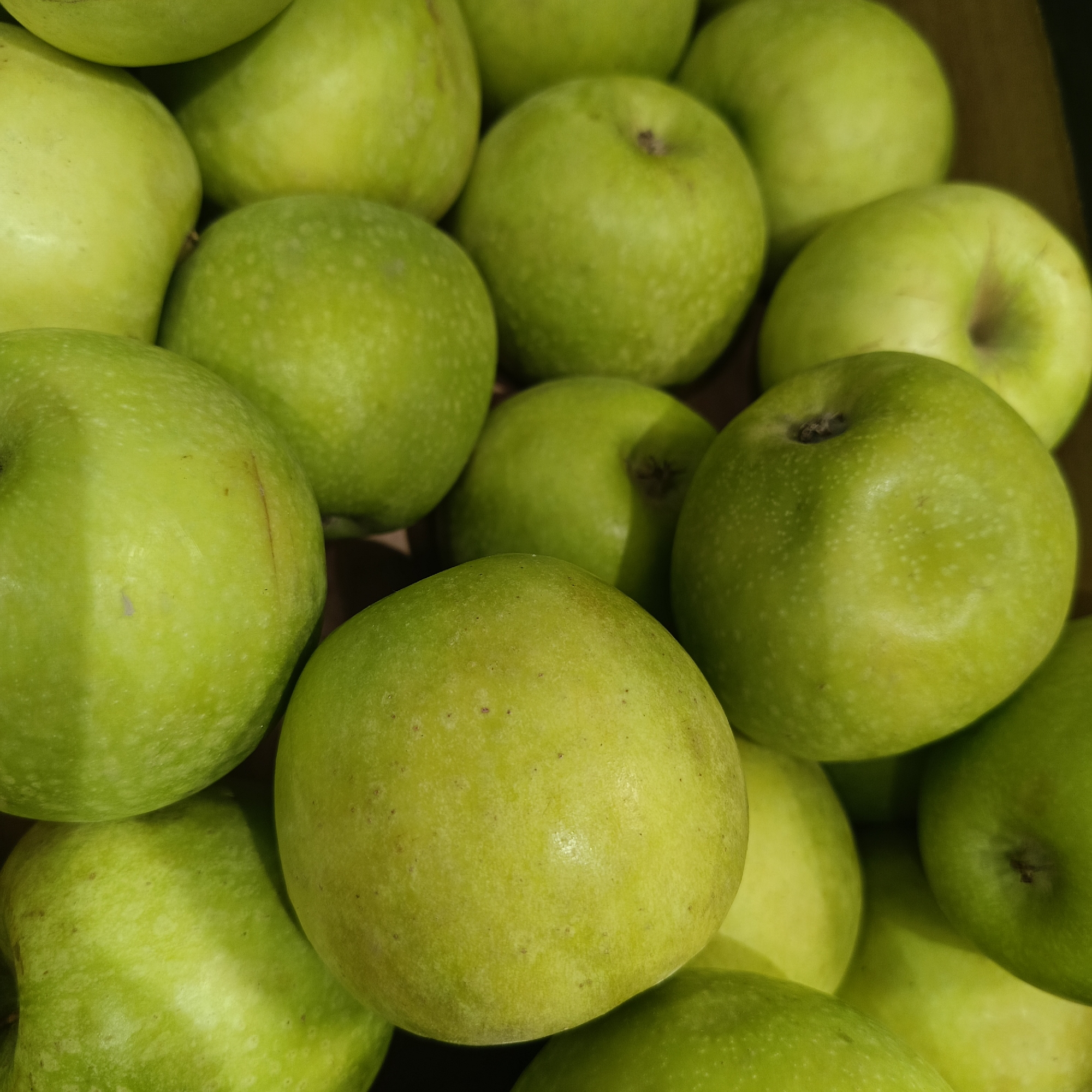 The image shows a close-up of a pile of green apples. The apples vary slightly in shade and have a mottled appearance. Some have stems or stem remnants visible. The background is indistinct. The location in the city of Марковский, Russia is not apparent from the visual information.