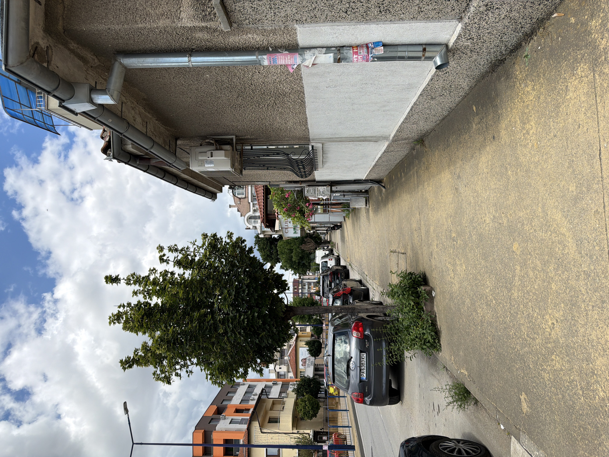 The image shows a street scene in Pazardzhik, Bulgaria. On the right is a light-colored building with a textured facade and a downspout. In front of the building is a concrete sidewalk. On the left side of the image is a street with several parked cars, and a tree in the foreground. Beyond the cars and the tree, there are more buildings and a street light. The sky is cloudy.
