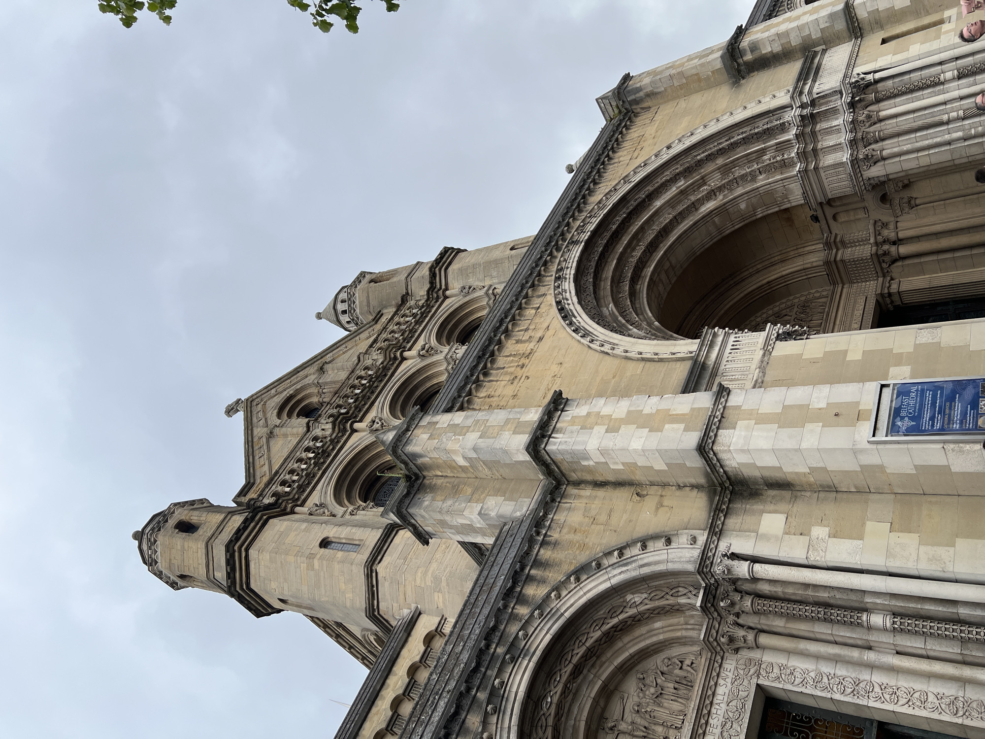 The image captures a low-angle view of Belfast Cathedral in Belfast City District, United Kingdom. The imposing stone facade of the cathedral dominates the frame, with its intricate carvings and architectural details highlighted under an overcast sky. Several arched entrances and decorative stonework are visible. A blue sign with white text, likely providing information about the cathedral, is mounted on the wall to the right of a side entrance. The sky is a uniform grey, suggesting cloudy weather. A few leaves from a tree are visible in the upper right corner of the image. There are no people prominently featured in the image, though a few individuals might be visible in the lower portion of the frame if zoomed in. The overall scene conveys a sense of grandeur and historical significance.
