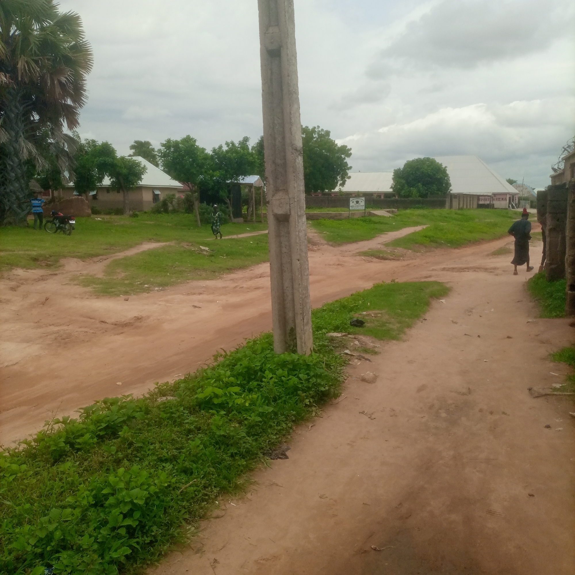 In an outdoor scene in an unidentifiable urban area, a dirt road with patches of grass and greenery runs through the frame. The sky is overcast with clouds. In the foreground, a large concrete utility pole stands prominently. Further back, several buildings with light-colored roofs are visible, surrounded by trees. To the left, two people and a motorcycle are near a palm tree. To the right, a person wearing a dark garment and a red hat walks away from the camera along the dirt road. A few other structures and people are sparsely scattered throughout the background.