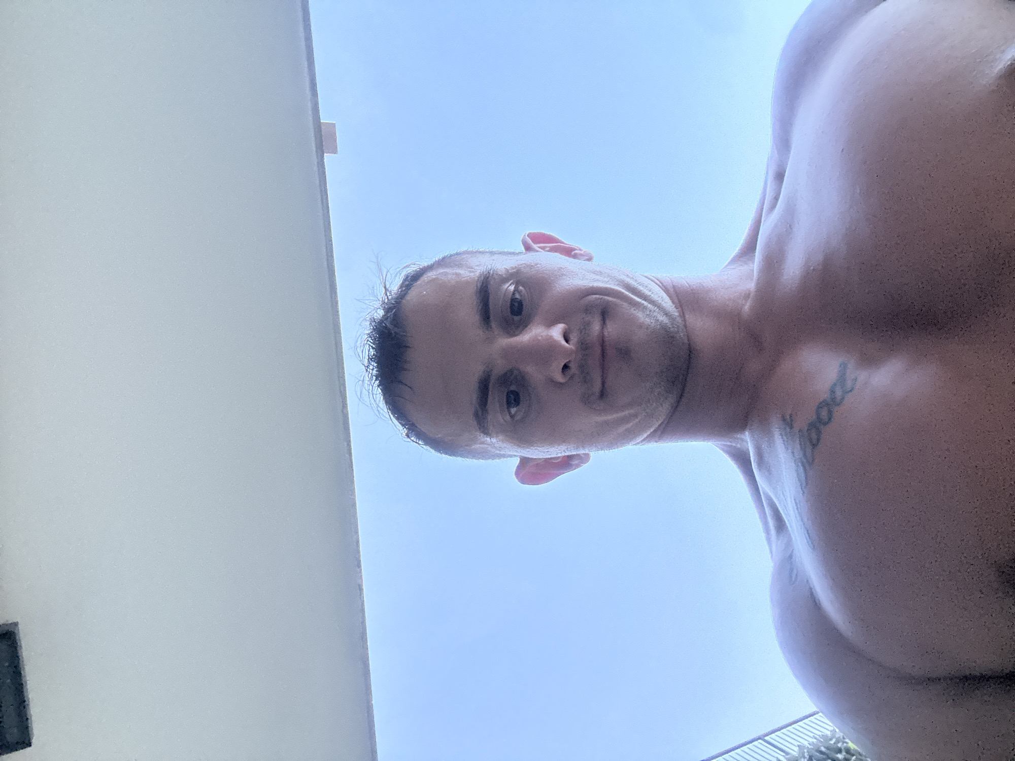 A low-angle, wide shot shows a man with a muscular physique looking up at the camera. The man has short, dark hair and is sweating. He has a tattoo on his chest. The background is a bright blue sky with some clouds. To the left of the man, a white structure, possibly a wall or building, takes up a significant portion of the frame. The bottom right corner of the image shows a glimpse of a fence and some greenery, suggesting an outdoor setting. The lighting suggests it is daytime, possibly midday given the brightness of the sky. The man's expression is neutral, and the overall mood is casual and possibly recreational, given the outdoor setting and the man's attire, or lack thereof. The location is identified as Finestrat, Spain.