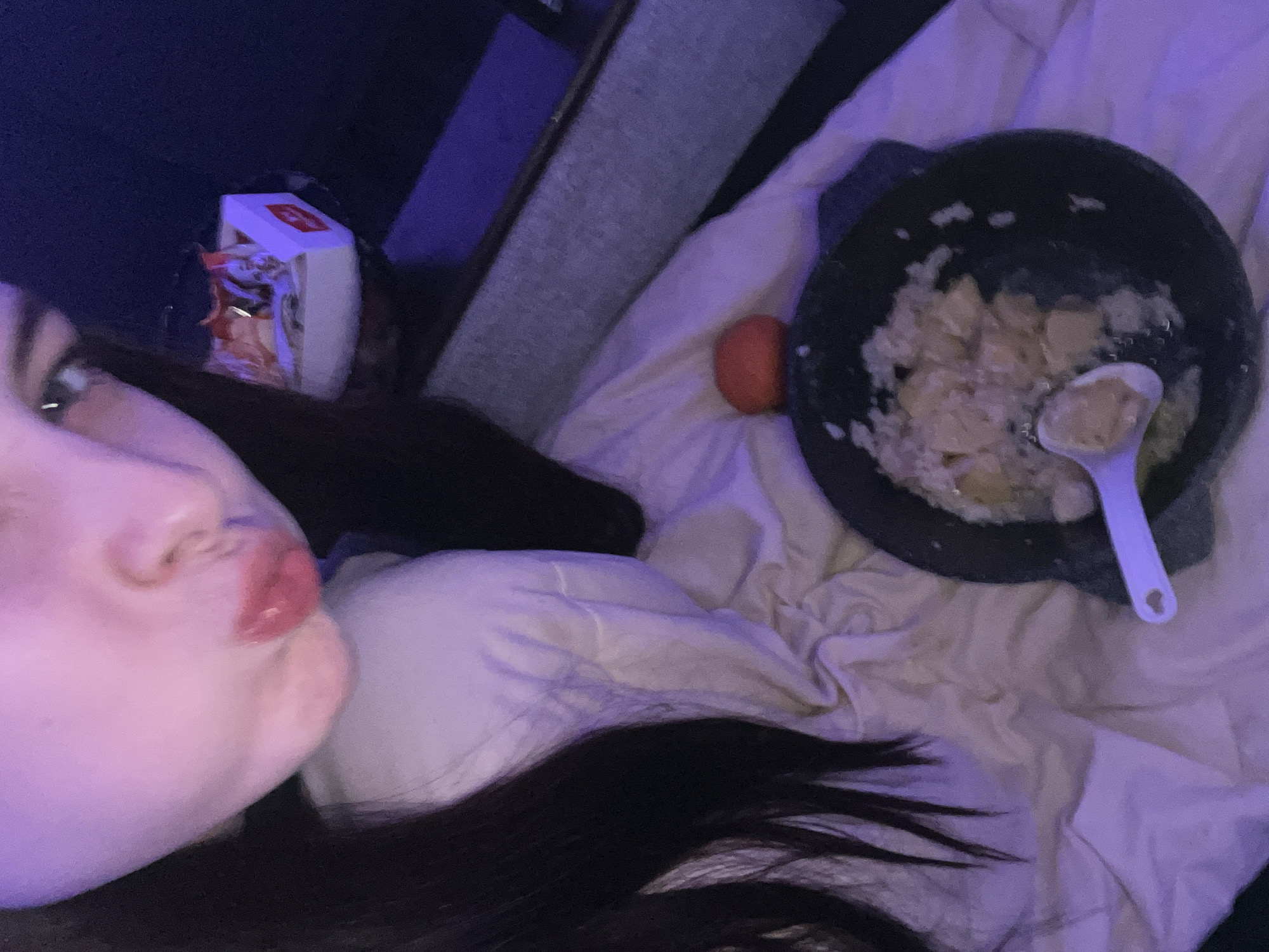 A person is lying in a bed, with a bowl of food and a spoon next to them. There is also an orange and a box of what appears to be food or snacks. The environment seems to be a bedroom setting. There is no evidence to indicate that the location is in the city of Saint Petersburg, Russia.