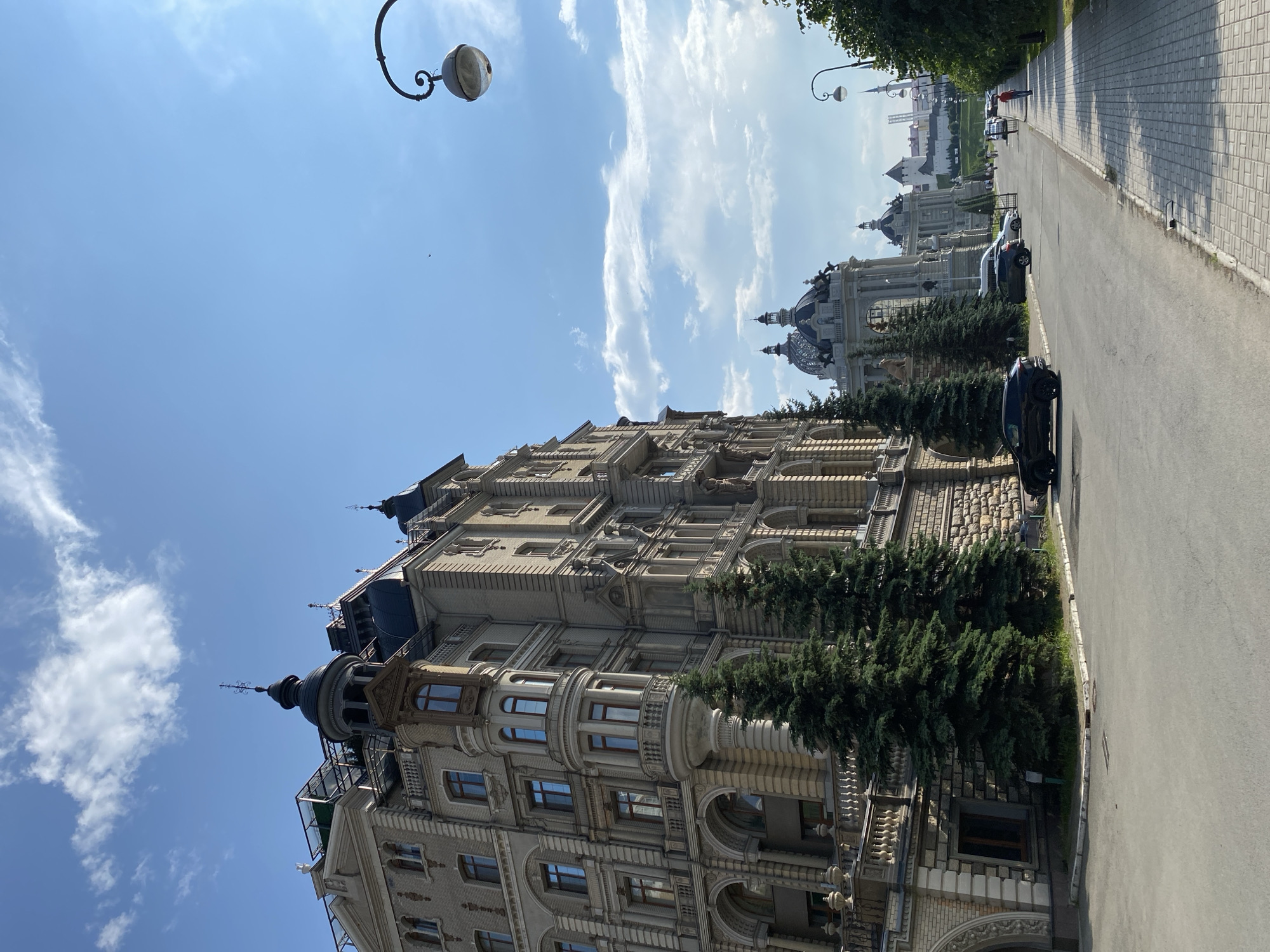 In the city of Bogorodskoe, Russia, on a clear day with scattered clouds, an ornate building with intricate architectural details stands tall on the left side of the frame. The building features multiple stories, bay windows, decorative moldings, and a dark, pointed roof. Several evergreen trees are planted in front of the building. A paved road runs alongside the building, with a sidewalk on the right. Several cars are parked along the road, and a few pedestrians are visible walking on the sidewalk. In the background, more buildings with domed roofs can be seen, along with a distant white tower. A street lamp with a curved arm extends into the sky from the right side of the image.