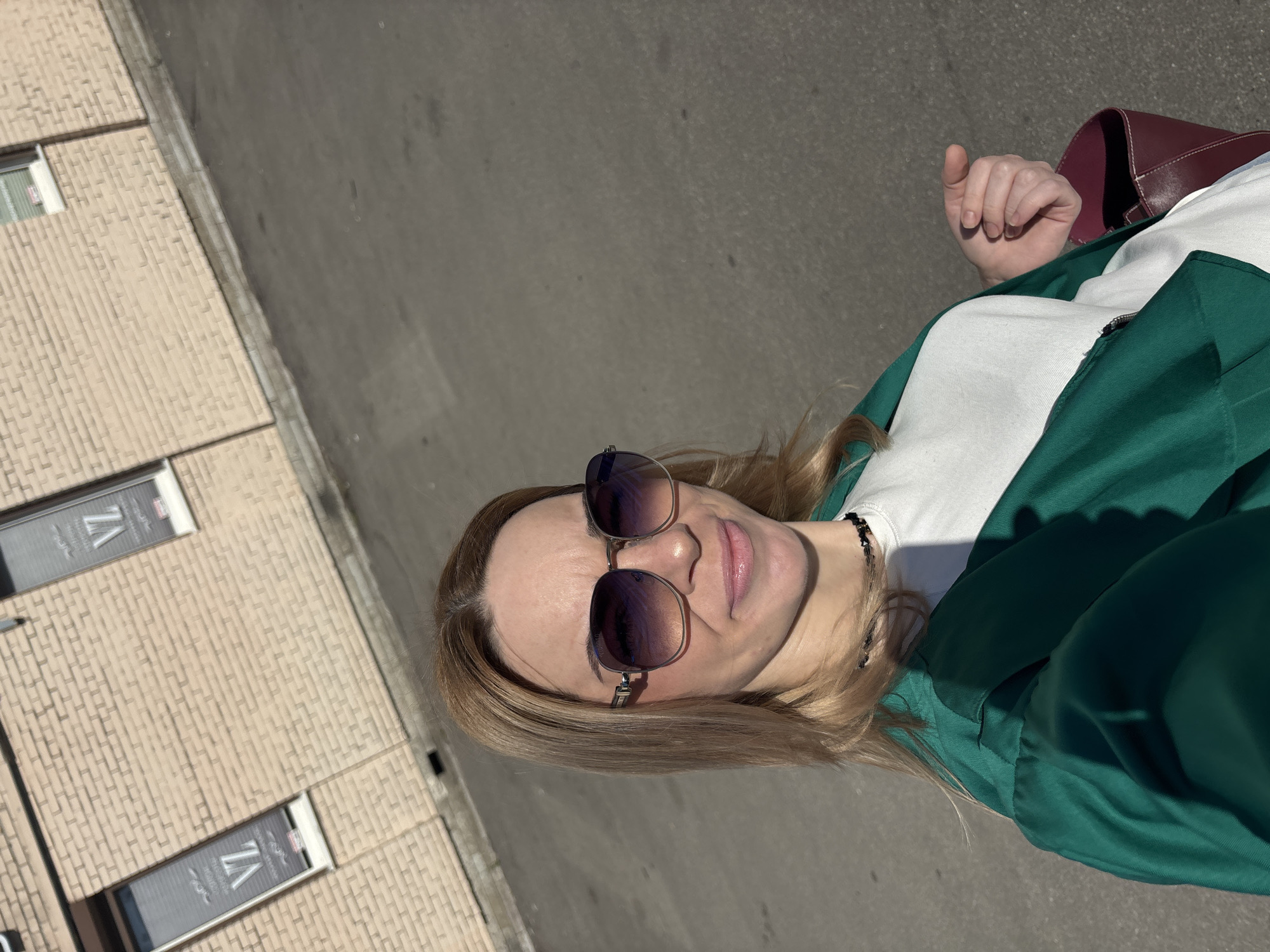 The image displays a woman posing outdoors on a bright, sunny day, captured from the chest up. Her head is tilted slightly, and she looks directly at the viewer with a subtle smile. She has light brown hair and wears dark-lensed sunglasses with metallic frames. Her outfit consists of a white t-shirt layered under a dark green jacket or hoodie, which is partially unzipped. A dark, possibly beaded necklace is visible around her neck. Her right hand is seen, holding a portion of a red or burgundy bag.
The background shows a paved surface, likely asphalt or concrete, on the right side of the frame. To the left, there is a building constructed with light-colored, possibly brick or textured siding. Several windows are visible on the building. Upon closer inspection of the windows, there are signs featuring stylized text, with "7A" being clearly discernible along with other less distinct lettering.
The presence of strong shadows indicates bright daylight and clear weather. The scene suggests an urban or suburban environment, possibly a street or sidewalk alongside a building.