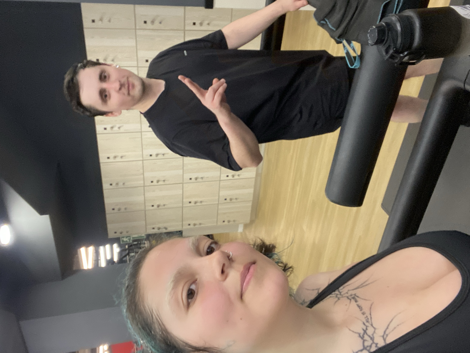 Two individuals are visible in the image, seemingly at a gym. One person, presumably taking the selfie, is a woman with a nose ring and a tattoo on her chest. The second person is a man behind her, making a peace sign with his hand. The background appears to be a gym setting, with lockers and exercise equipment visible. The location in the city of Gijón/Xixón, Spain, cannot be determined from the image.