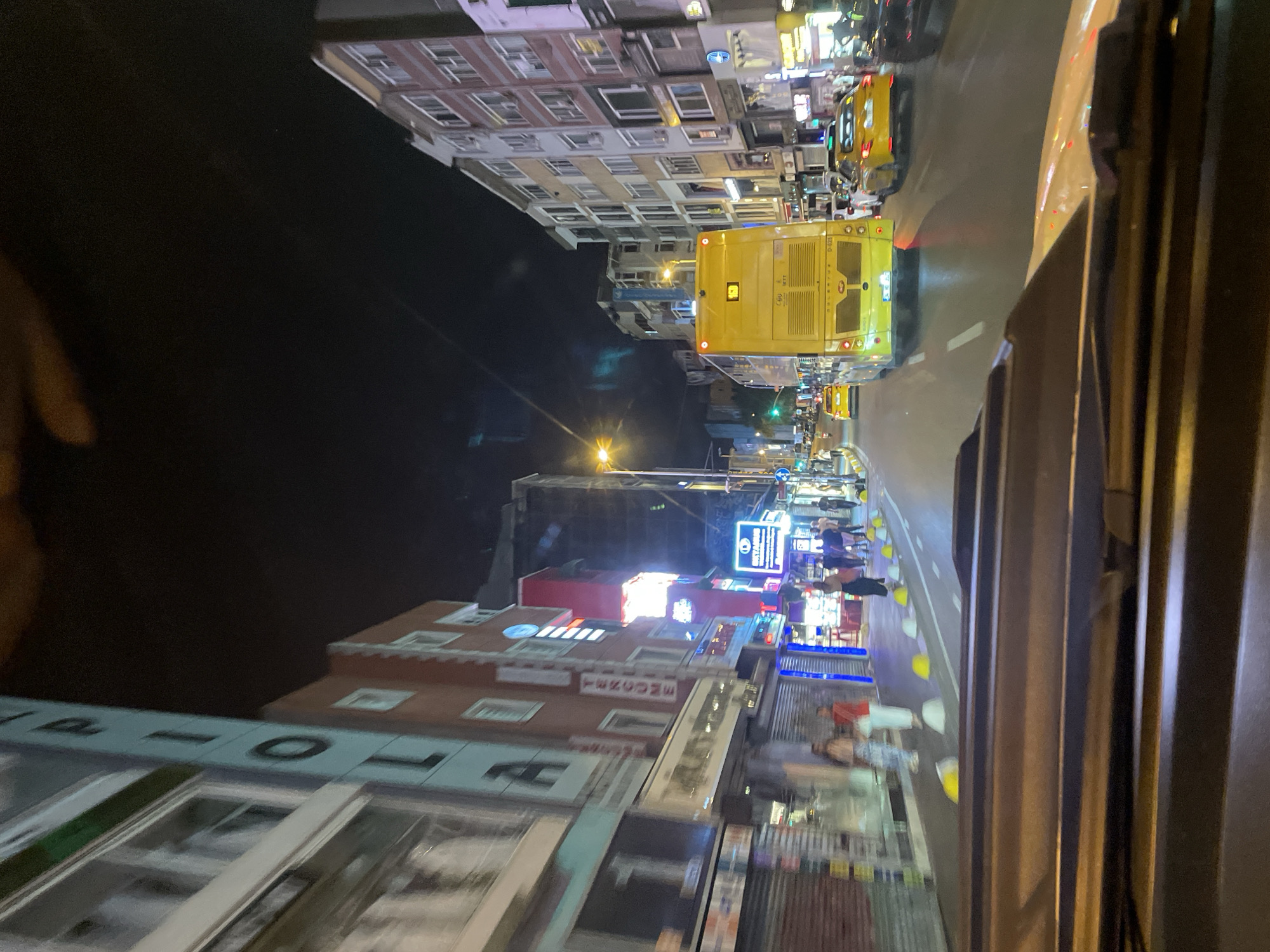 The image captures a night scene on a city street in Kadıköy, Turkey. The perspective is from inside a vehicle, looking forward through the windshield. A yellow bus is the prominent subject in the foreground, with its rear end facing the camera. The bus displays text and a license plate. Other vehicles, including yellow taxis and a black car, are also visible on the road.
The street is lined with multi-story buildings, some of which have illuminated signs and windows. The facades of these buildings are varied in color and architectural style, suggesting a commercial or residential area. Pedestrians are present on the sidewalks, with some individuals appearing to walk or stand in groups. Streetlights and building lights illuminate the scene, casting reflections on the wet or polished road surface.
Yellow and white bollards are strategically placed along the edge of the road. The overall atmosphere is bustling, characteristic of a lively urban environment at night. The sky is dark, indicating nighttime.
