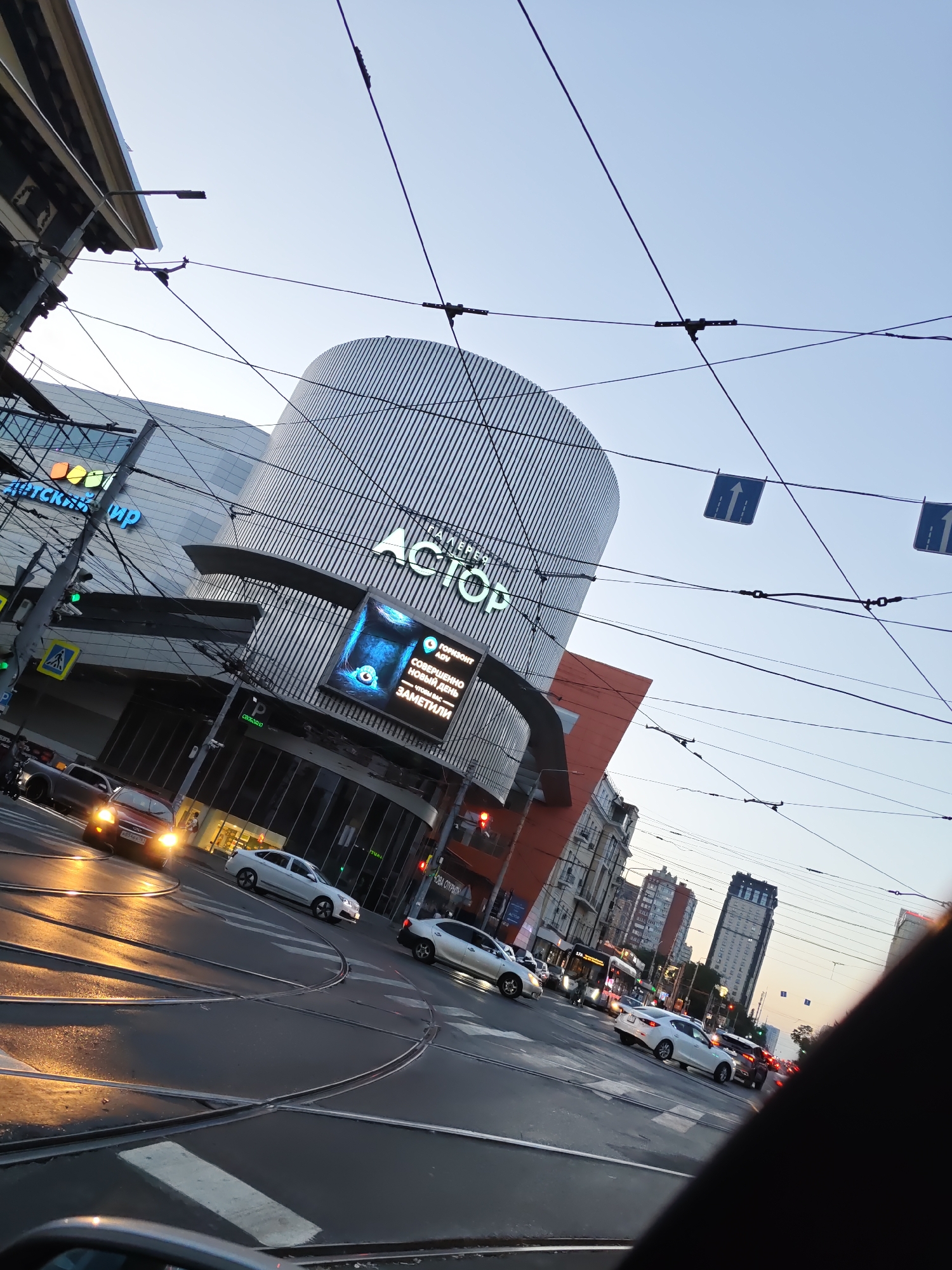 The image captures a street scene in Rostov-on-Don, Russia, likely during the evening or early morning given the lighting and illuminated signs. The focal point is the modern, cylindrical building of the "Gulliver" shopping center, prominently featuring its name in illuminated white lettering. Adjacent to it is another large building, possibly part of the same complex, with a facade of vertical metal louvers.
The street is a multi-lane road with visible tram tracks. Several vehicles, including cars and a bus, are present on the road, with some traffic lights visible. The wet asphalt suggests recent rain. Overhead, a complex network of power lines for trams crisscrosses the sky.
In the foreground, the edge of a vehicle's interior is visible, suggesting the photo was taken from inside a car. The overall atmosphere is urban and dynamic, with a blend of modern architecture and active city life.