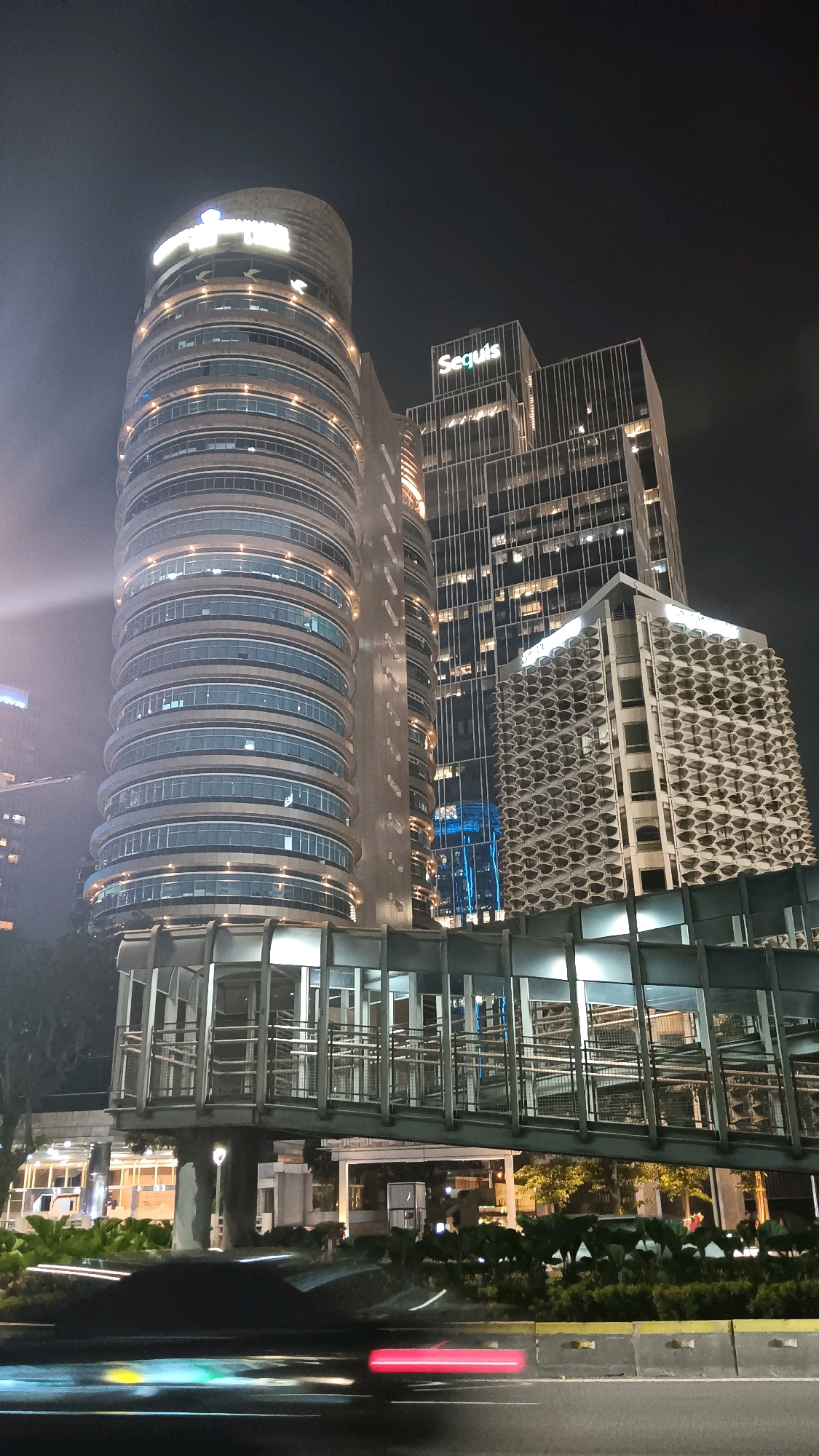 An outdoor, low-angle, nighttime, vertical photograph shows the Wisma 46 building in Central Jakarta, Indonesia. The building is a tall, cylindrical skyscraper with a ribbed facade, illuminated from within by rows of windows. To the right of the Wisma 46 building, a rectangular skyscraper with a grid-like facade is also illuminated by internal lights. In front of these two buildings is a pedestrian bridge, with a metal frame and glass walls, supported by concrete pillars. The bridge spans across a street where the lights of passing vehicles are blurred by motion, creating streaks of red, yellow, and green. The street is lined with lush green foliage, and the sky above is dark and cloudy.