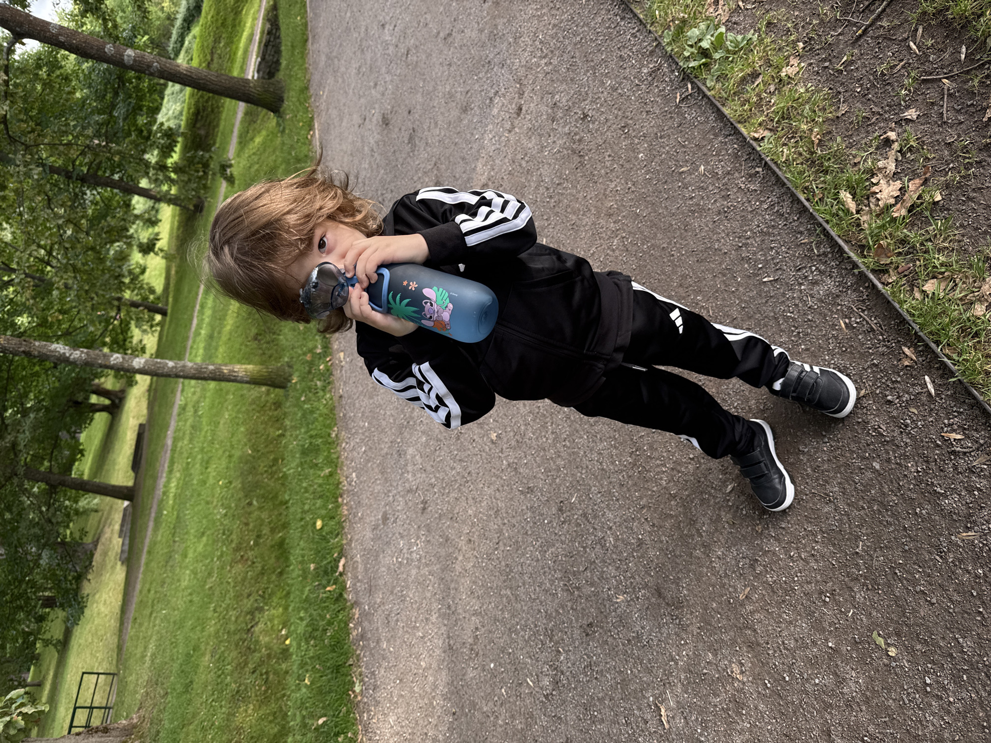 A young child with wavy brown hair is standing on a gravel path in a park, holding a blue water bottle up to their mouth and drinking from it. The child is wearing a black tracksuit with white stripes down the sleeves and legs, and black velcro sneakers. The park features a grassy slope, trees, and another path in the background. The setting appears to be a public park.