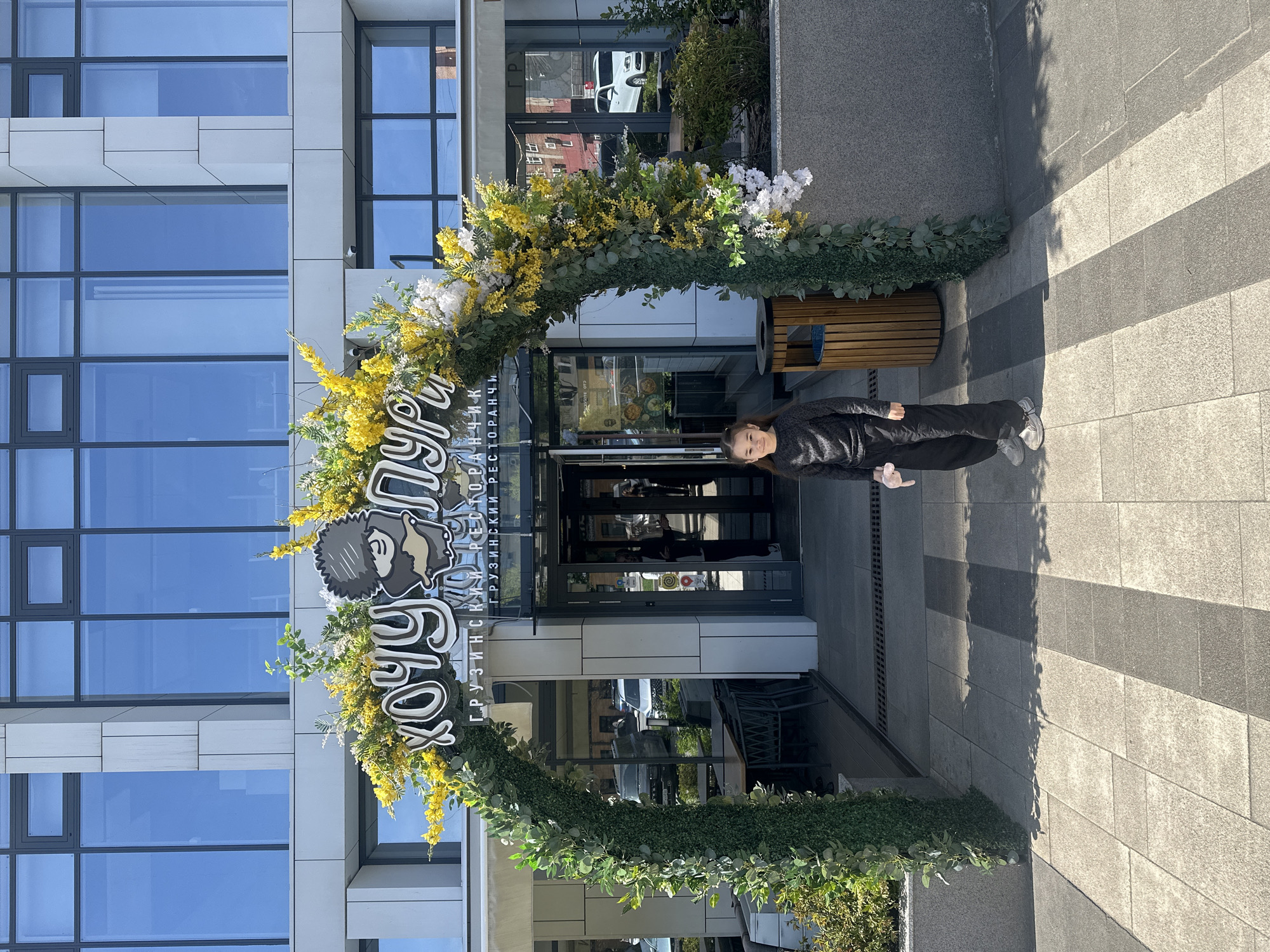 A woman stands in front of the entrance to the "Khokhlyandiya" Georgian restaurant in Izhevsk, Russia. The restaurant's entrance is decorated with a floral arch made of greenery, yellow, and white flowers. Above the entrance, the restaurant's sign is visible, featuring a cartoonish face and Cyrillic script. The building's facade includes modern gray panels and reflective windows. The woman is dressed in dark clothing and light-colored shoes. The ground is paved with gray stone. The lighting suggests a sunny day.