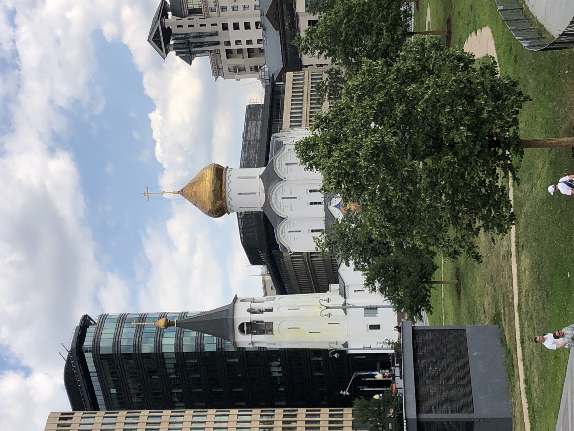 The image depicts a view of the Cathedral of Christ the Saviour in Moscow, Russia. In the foreground, a manicured lawn with trees and pathways is visible. Two men are walking on a path in the lower left corner. Behind the lawn, the white walls and golden dome of the cathedral are prominent, with a tall bell tower to its left. In the background, modern high-rise buildings, characteristic of Moscow's architecture, frame the historical structure. The sky is partly cloudy, with patches of blue. The scene appears to be captured on a bright day, suggesting it is likely daytime.