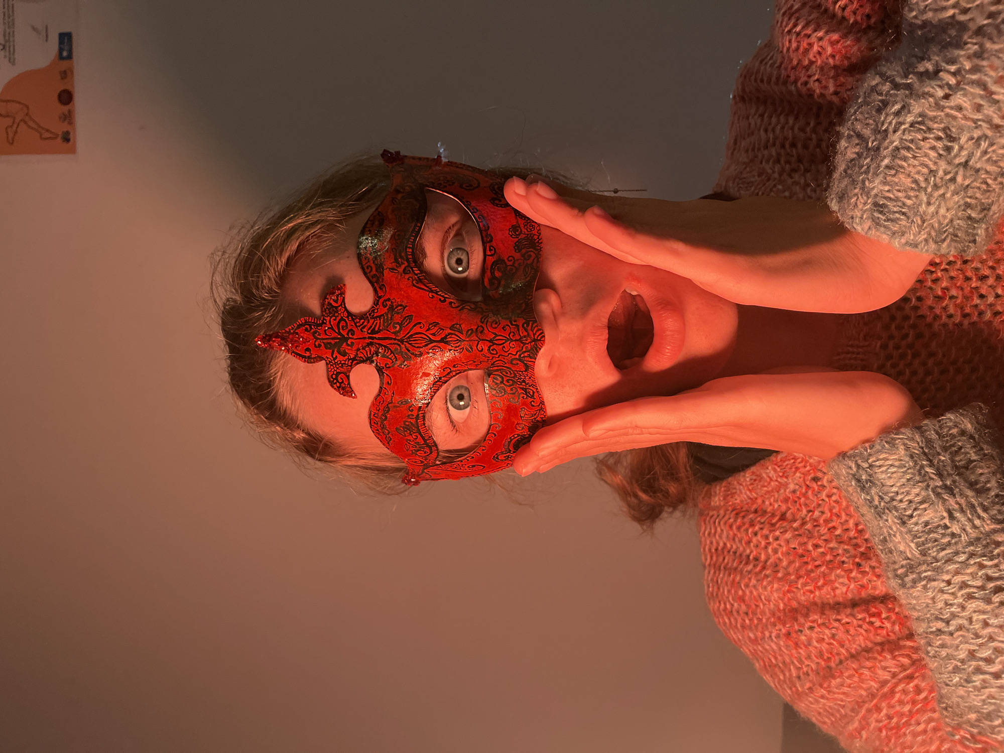 A person with light skin and light hair is wearing a red, ornate mask over the upper half of their face. The mask is designed with swirling patterns and covers the area around their eyes. Their eyes are wide and appear to be looking directly at the camera. Their mouth is open in an expression of surprise or shock. The person is holding their hands up to their face, framing the mask. They are wearing a chunky knit sweater in shades of red and grey. The background is a solid, light tan color, and a poster is visible in the top left. The image has an indoor, possibly studio-lit feel. There is no indication of Budva, Montenegro, in the picture.